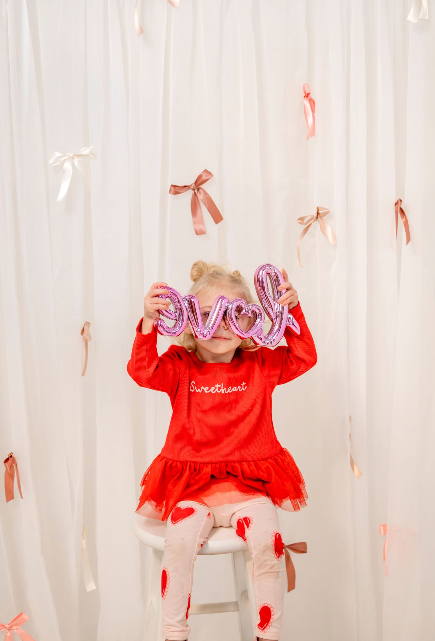Blonde girl in red Sweetheart dress holds love balloon letters over her eyes on a white stool.