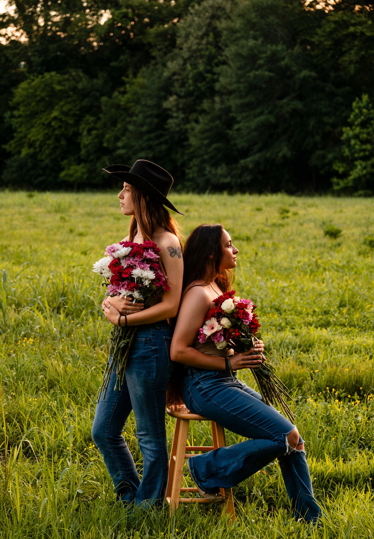 Two women in jeans hold colorful flower bouquets back-to-back on a stool in a lush green field at sunset.