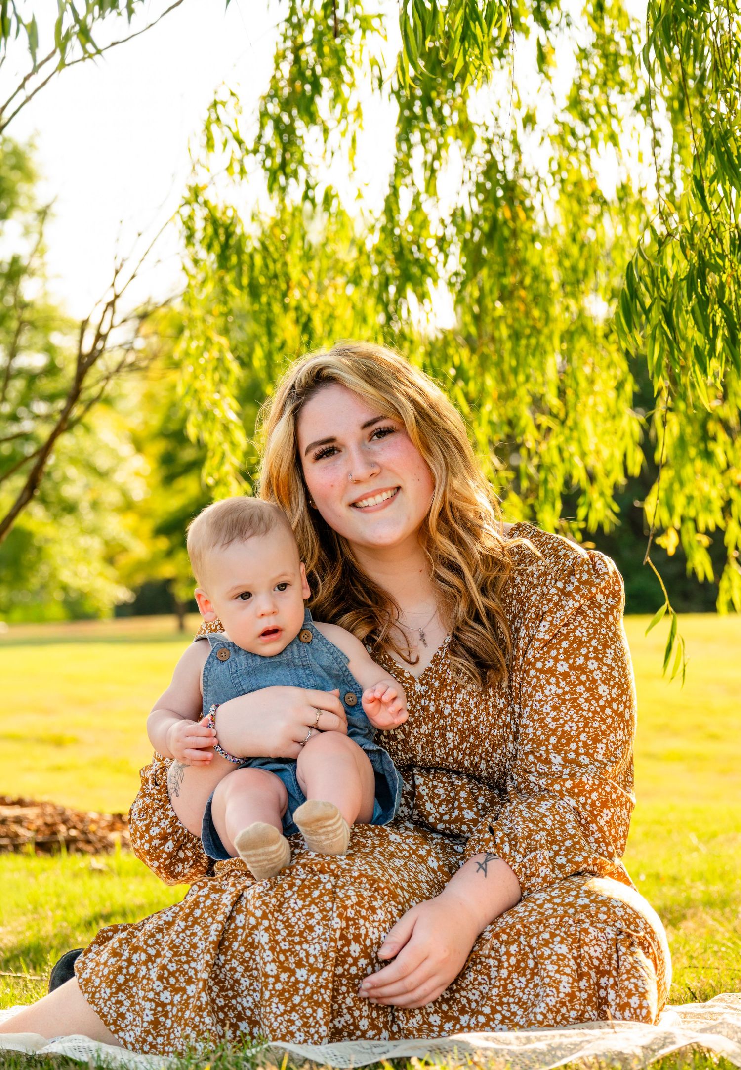 Young woman in floral dress smiling while holding a baby boy outdoors on sunny day near willow tree.