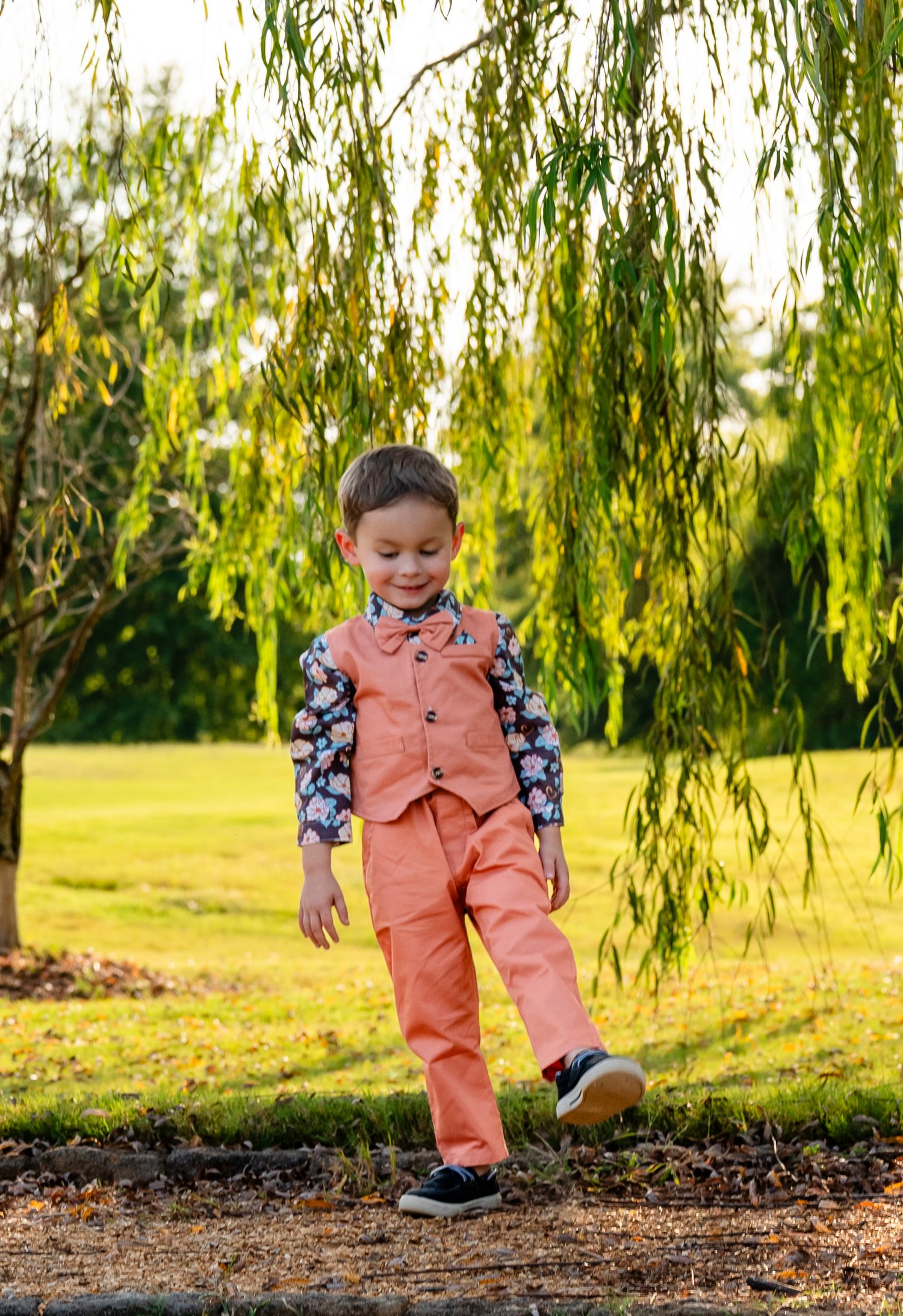 A young boy in a coral vest and floral shirt walks outdoors near a weeping willow tree.