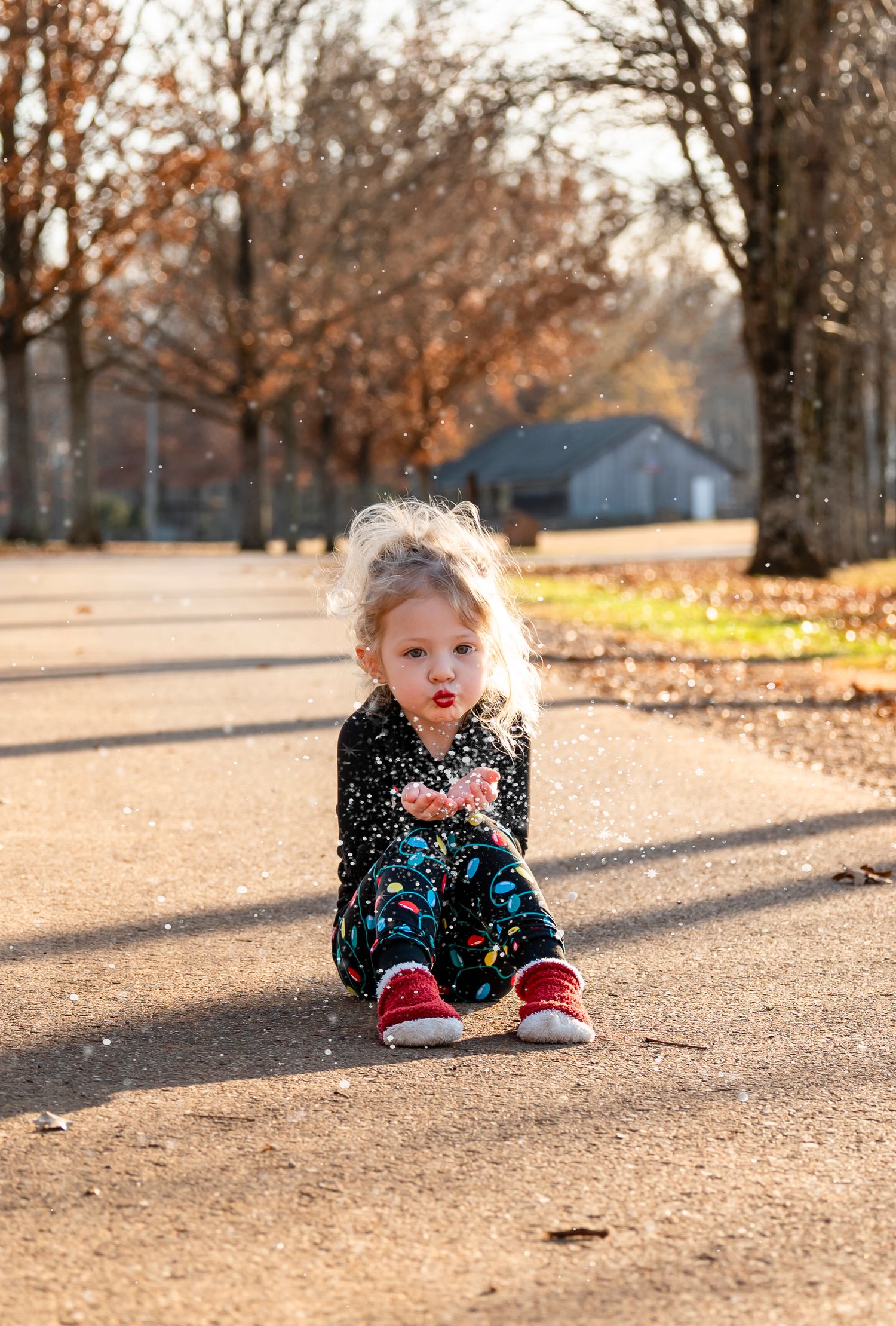 Adorable blonde toddler in Christmas pajamas sitting on sunny path blowing fake snow kisses.