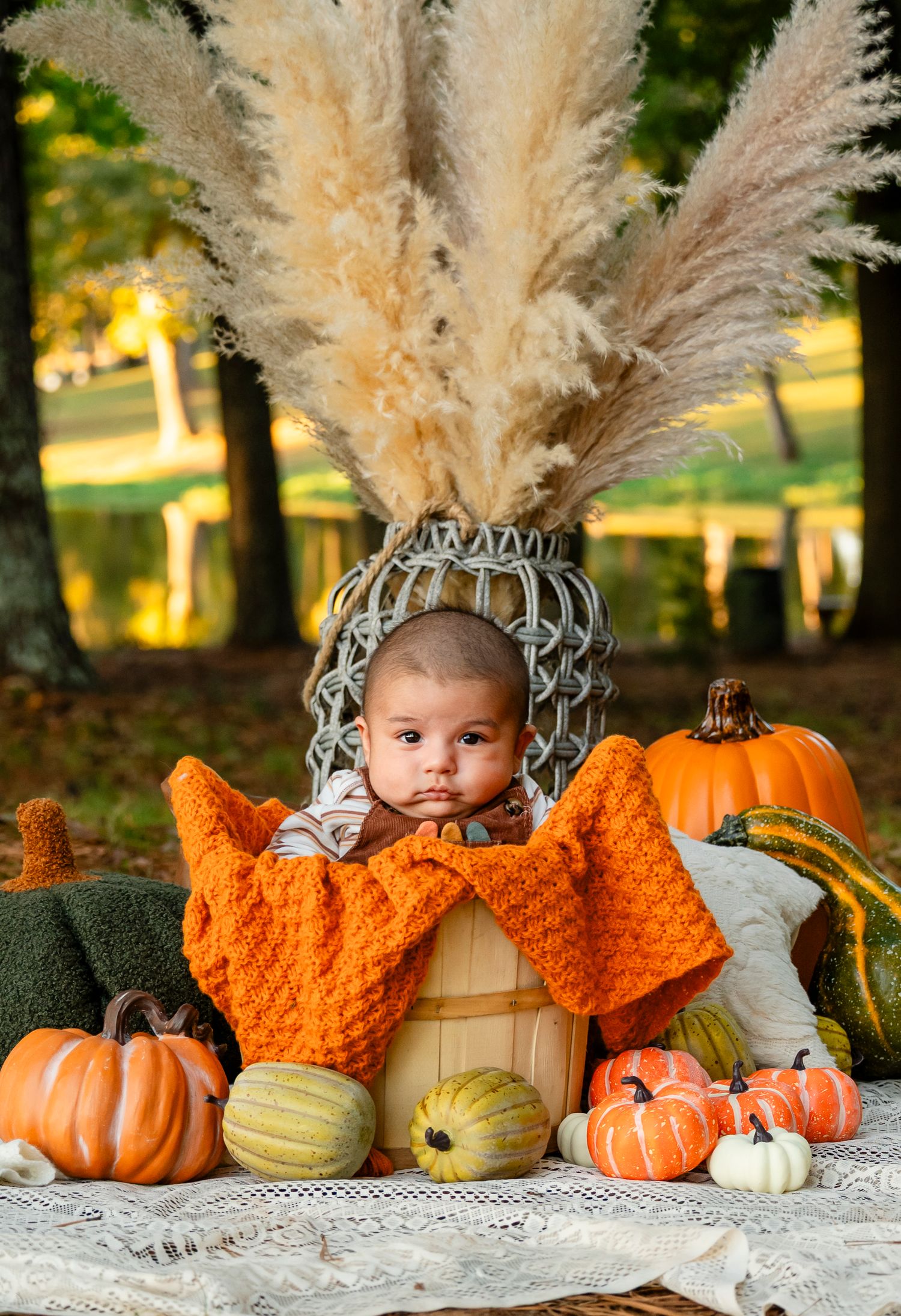 Baby nestled in wooden barrel with orange knit blanket surrounded by fall pumpkins outdoors.