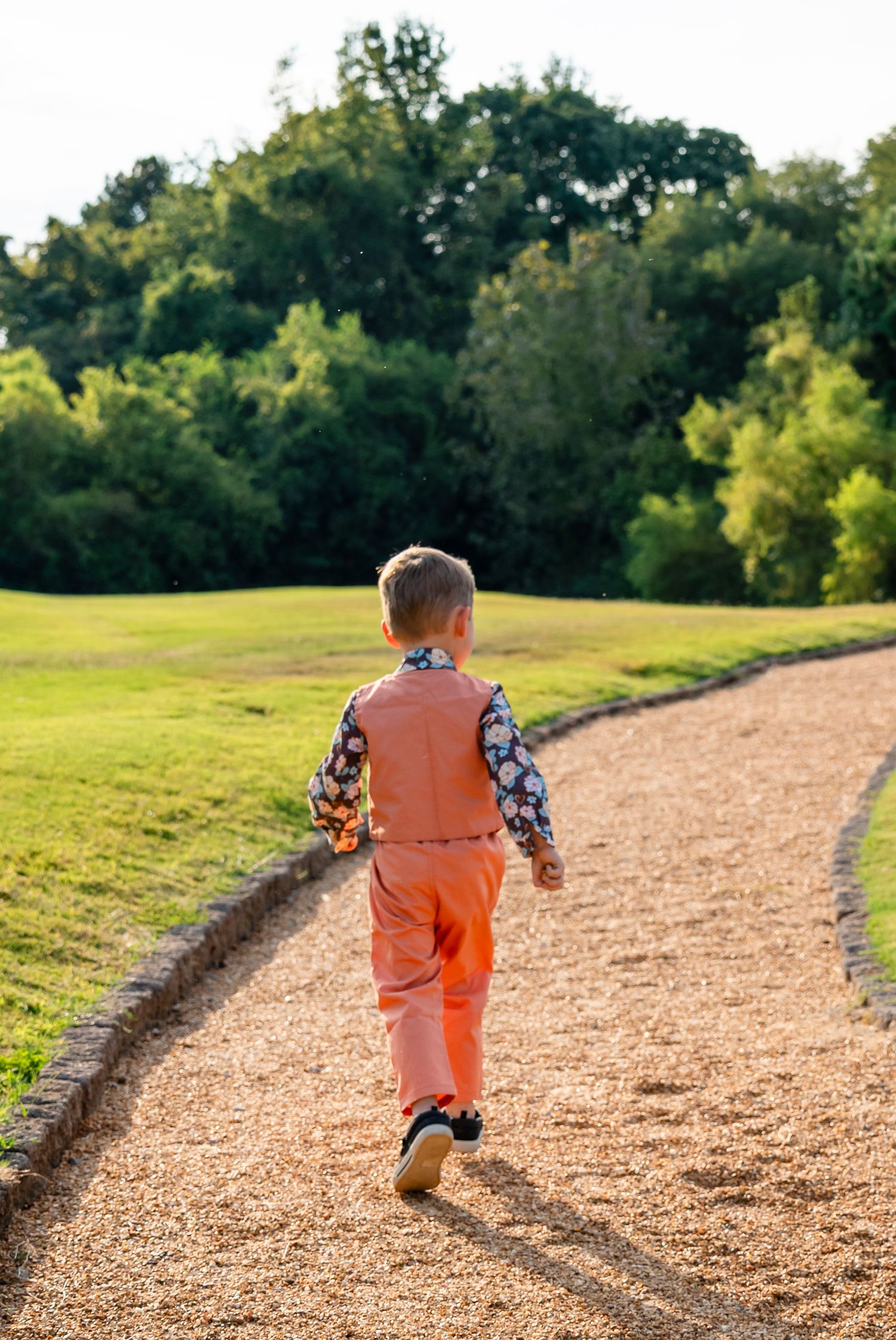 Young boy in orange pants and floral shirt walking away on a gravel path in a sunny green park.