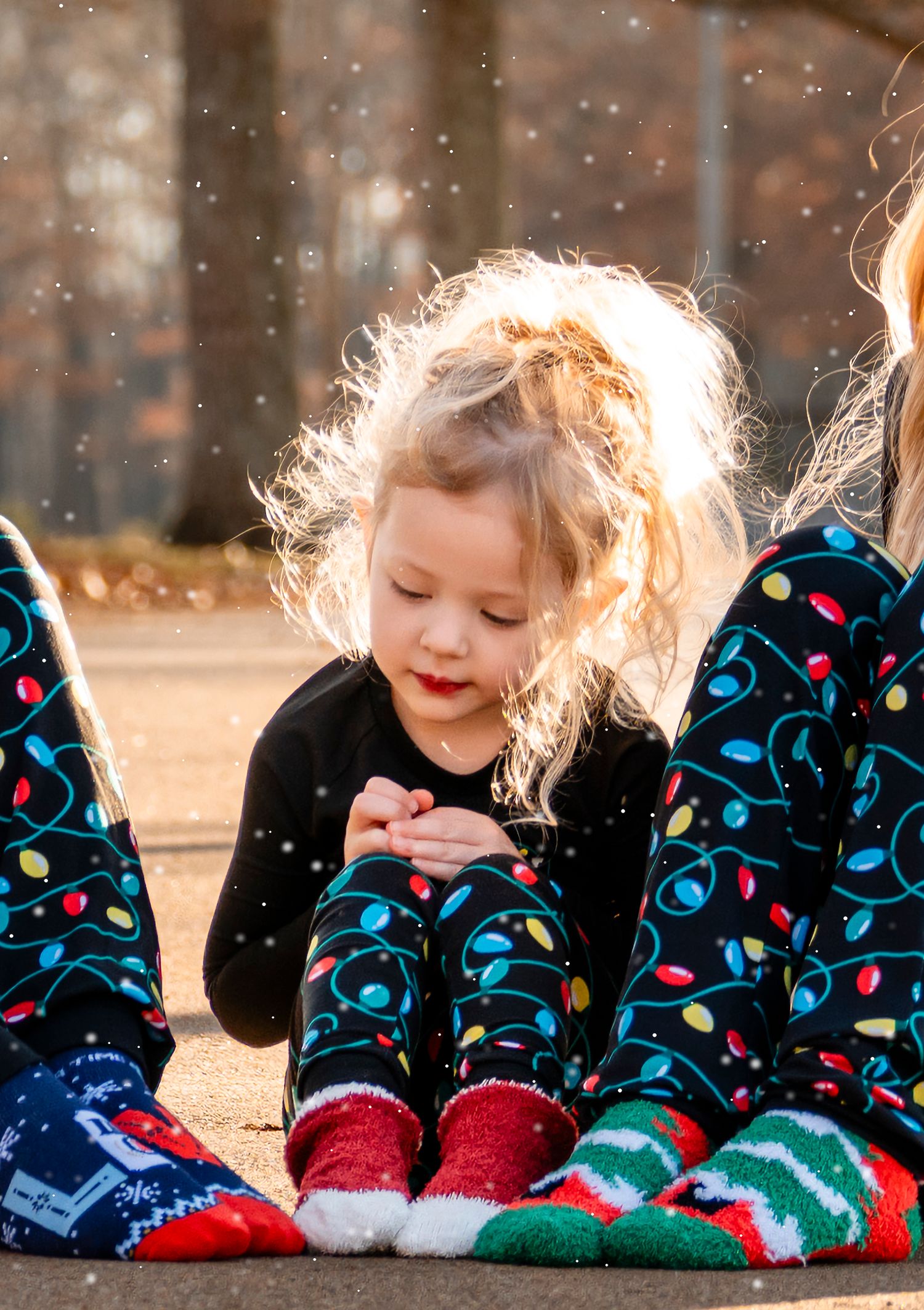 Close-up of blonde toddler in Christmas pajamas examining fake snow between two siblings outdoors.