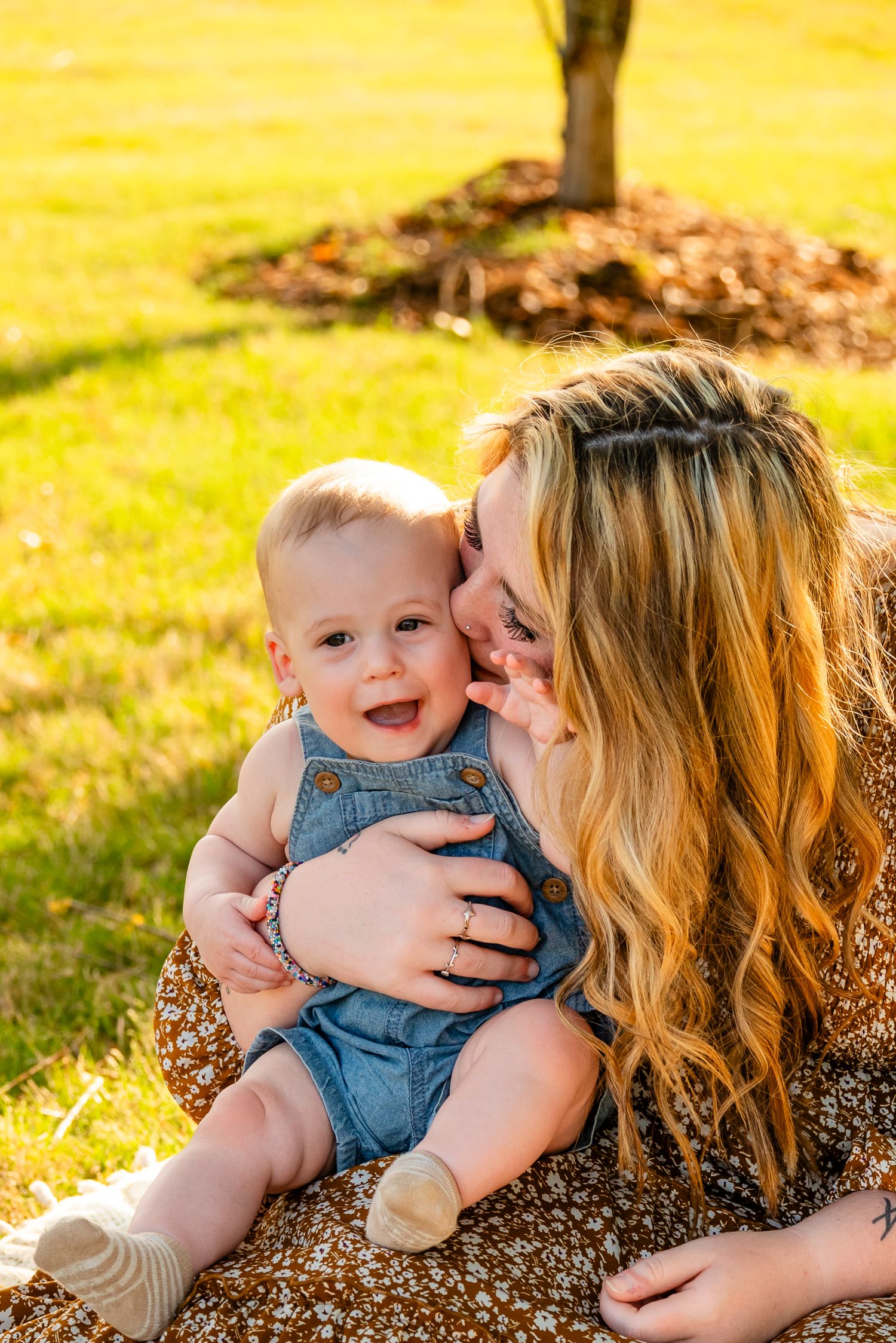 A joyful baby in denim overalls smiles while a blonde mother kisses their cheek outdoors on sunny grass.