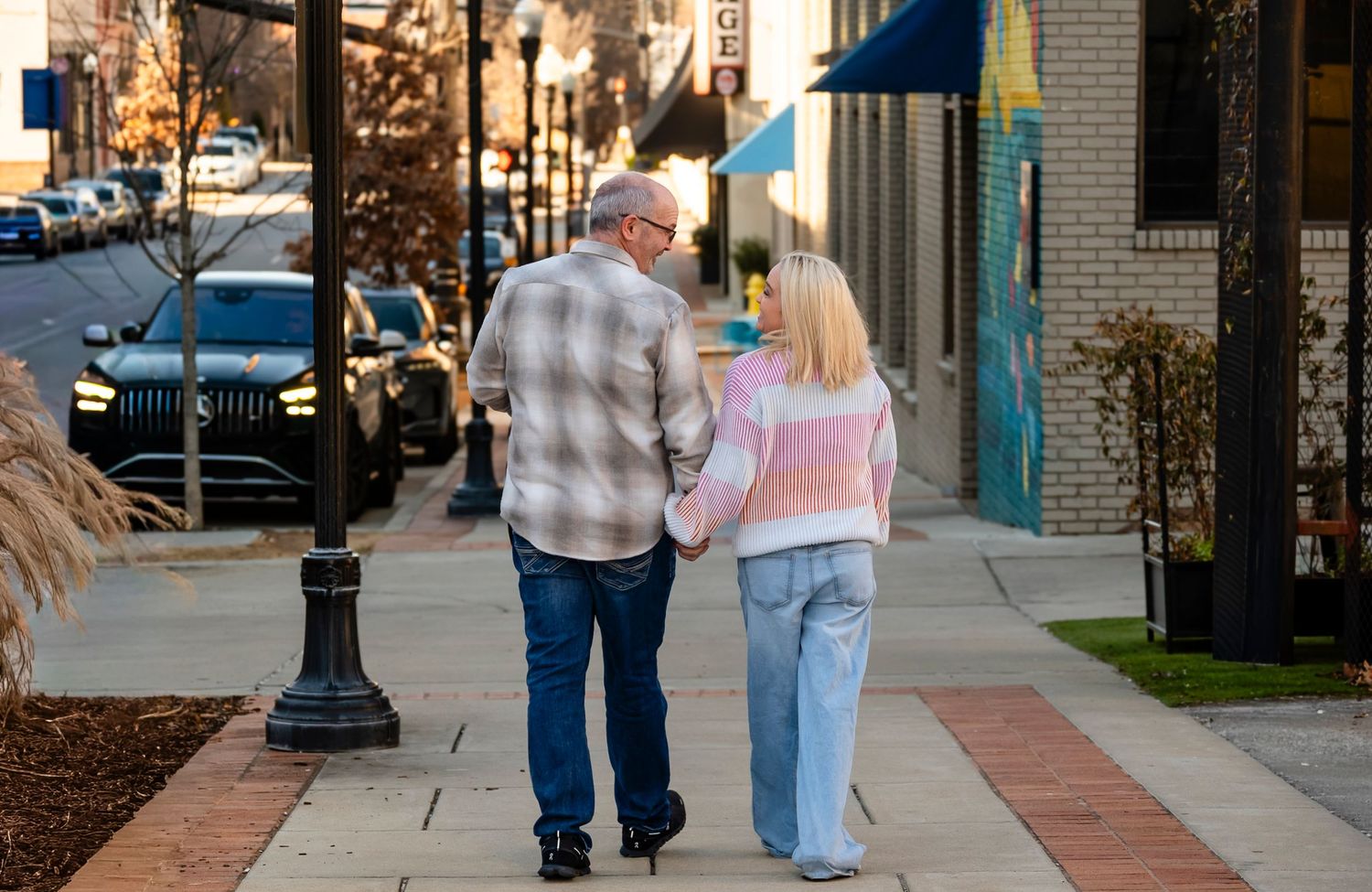 Older couple walking hand in hand down a sunny small-town sidewalk, viewed from behind.