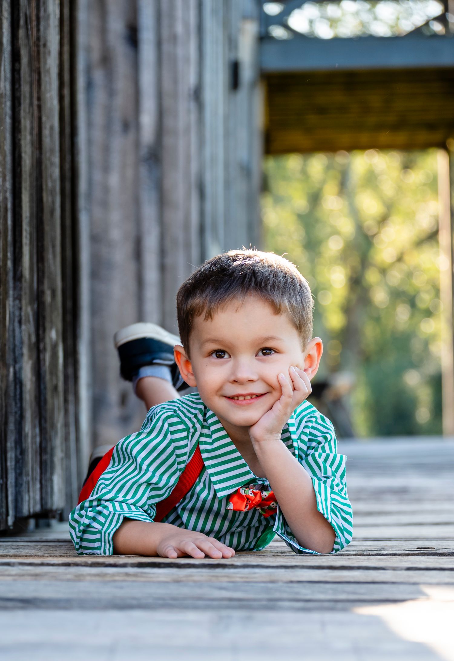 Smiling young boy in green striped shirt lying on rustic wooden deck with hand on cheek, outdoors.