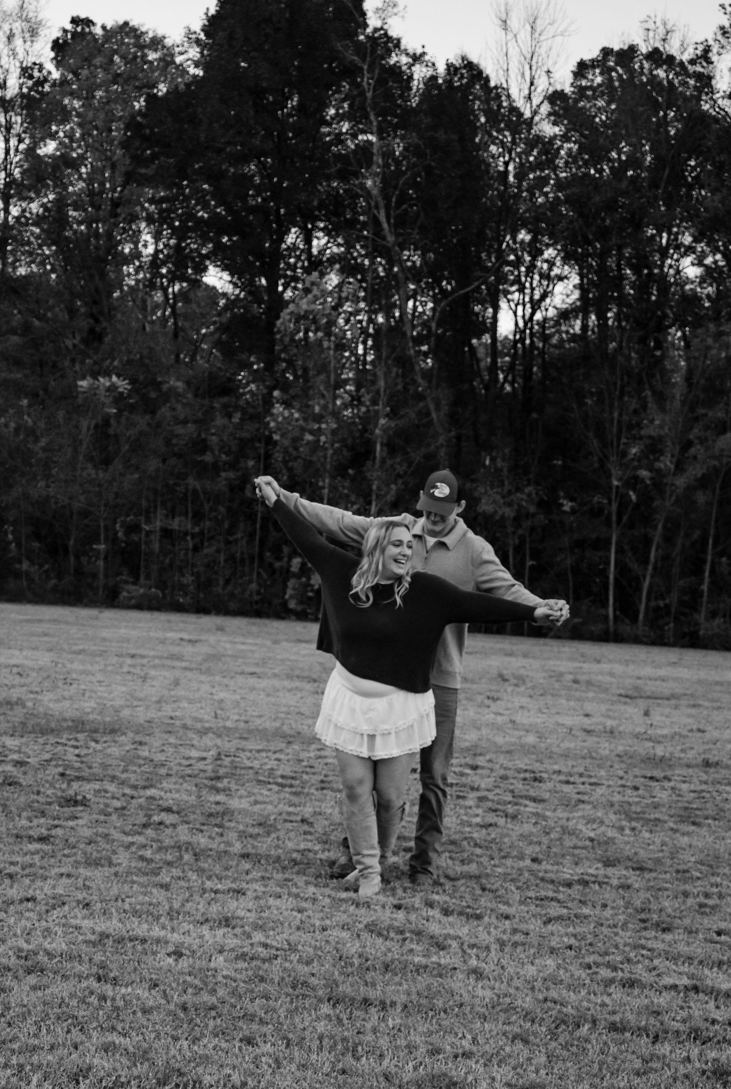 Black and white photo of a joyful couple dancing in an open field surrounded by trees.