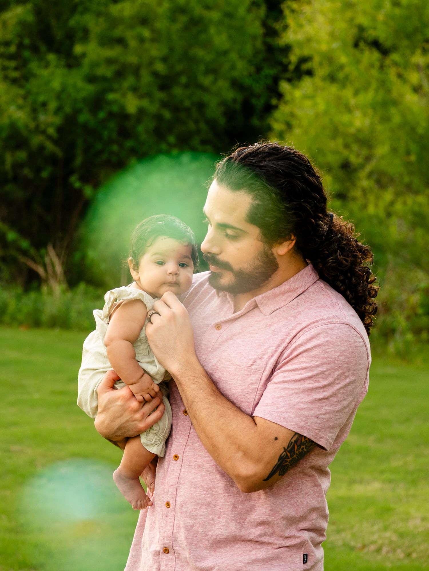 A man with long curly hair and a pink shirt tenderly holds a baby outdoors in a lush green park setting.