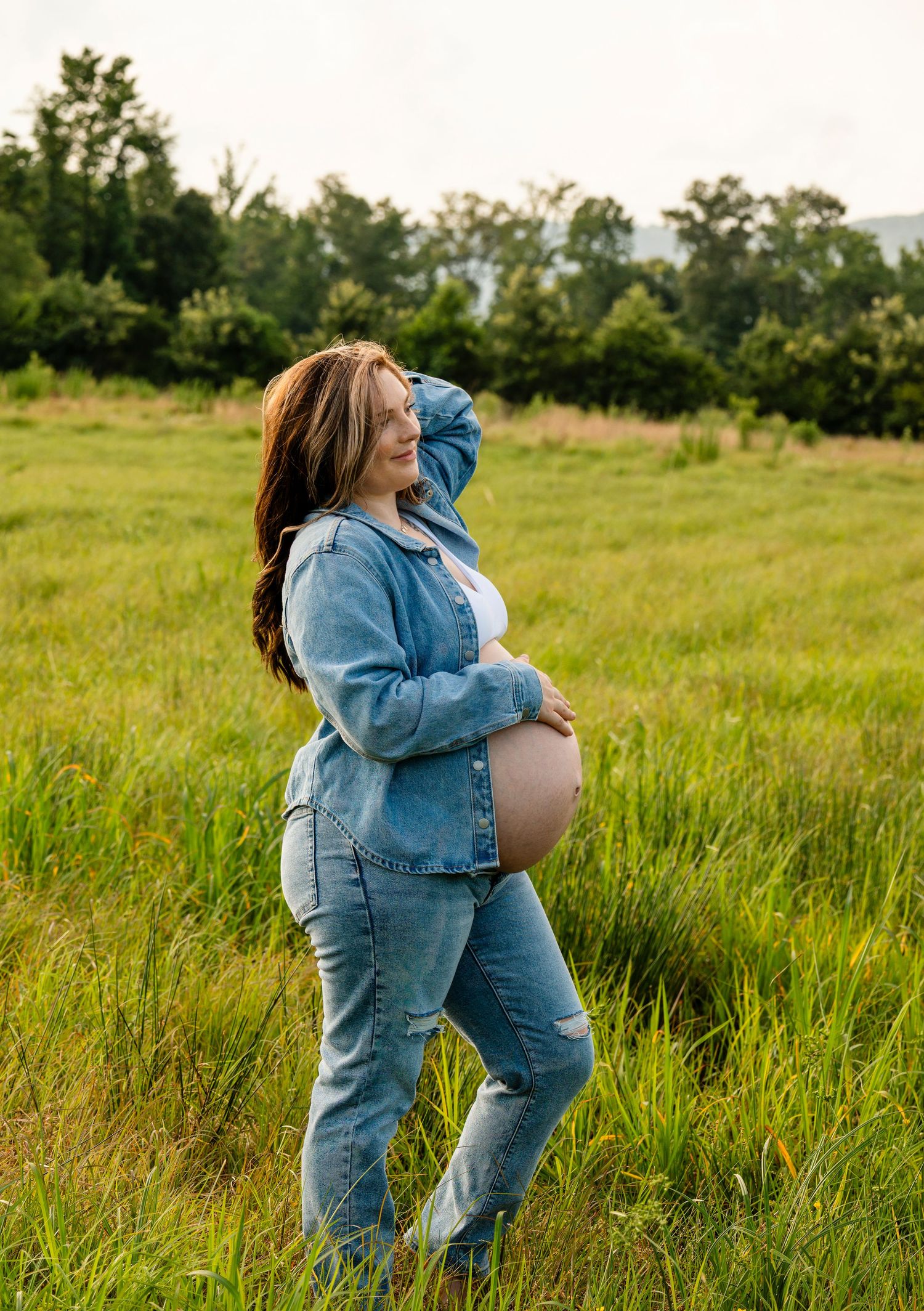 Pregnant woman in denim jacket and jeans standing in green field, cradling baby bump while smiling outdoors.