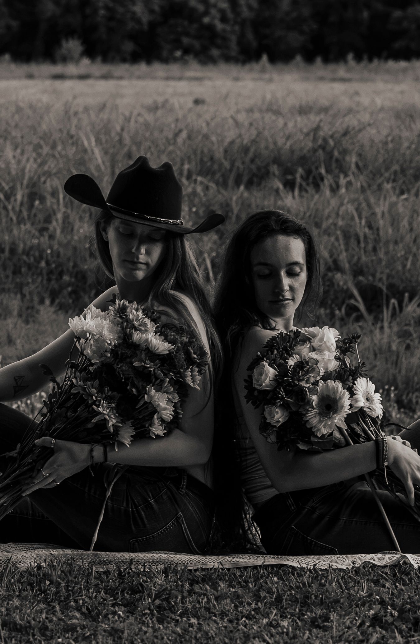Black and white photo of two women sitting back-to-back holding bouquets in a field, one in a cowboy hat.