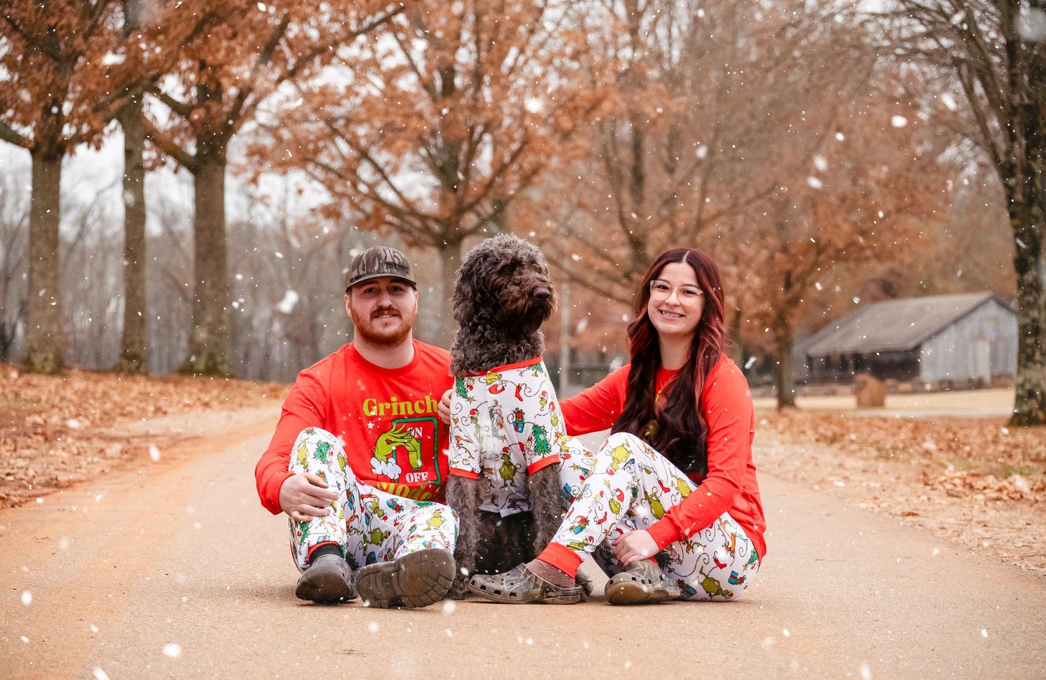 Couple and their curly dog sit together on a leaf-covered path surrounded by autumn trees.