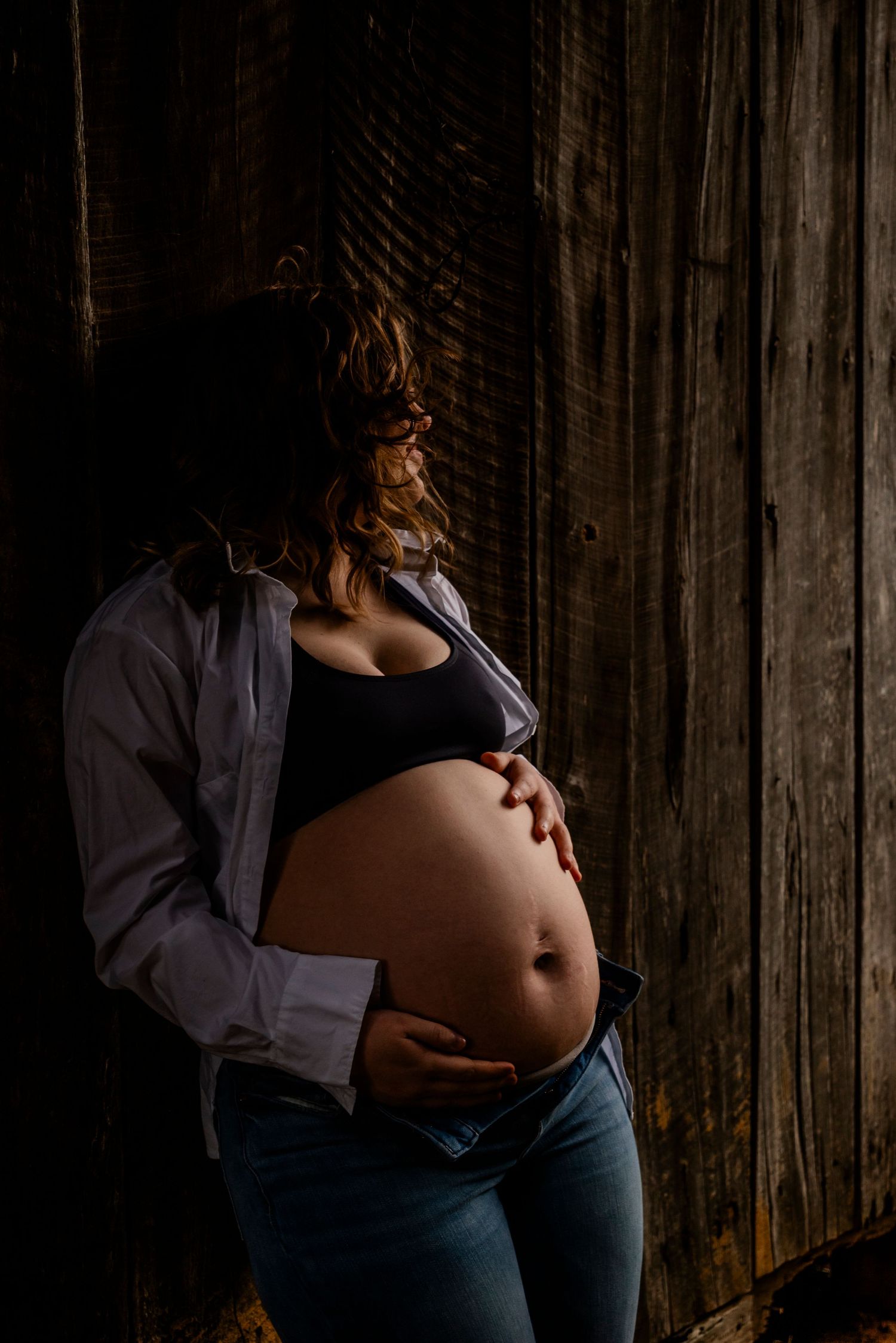 Pregnant woman in white shirt and jeans cradles bare baby bump against dark wooden barn wall, moody maternity portrait.