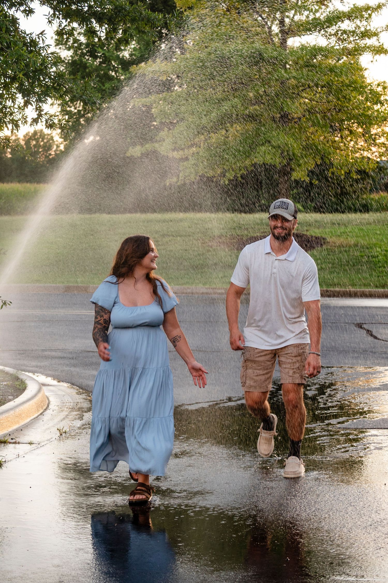 A couple walks and laughs together through a sprinkler spray on a wet road at golden hour.