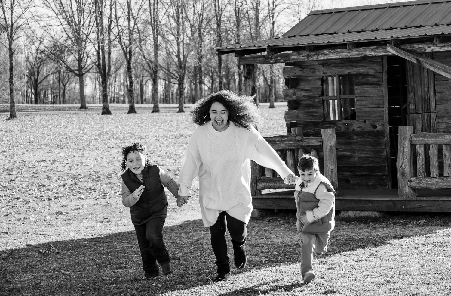 Black and white photo of a mother running and laughing with her two young sons near a rustic cabin.