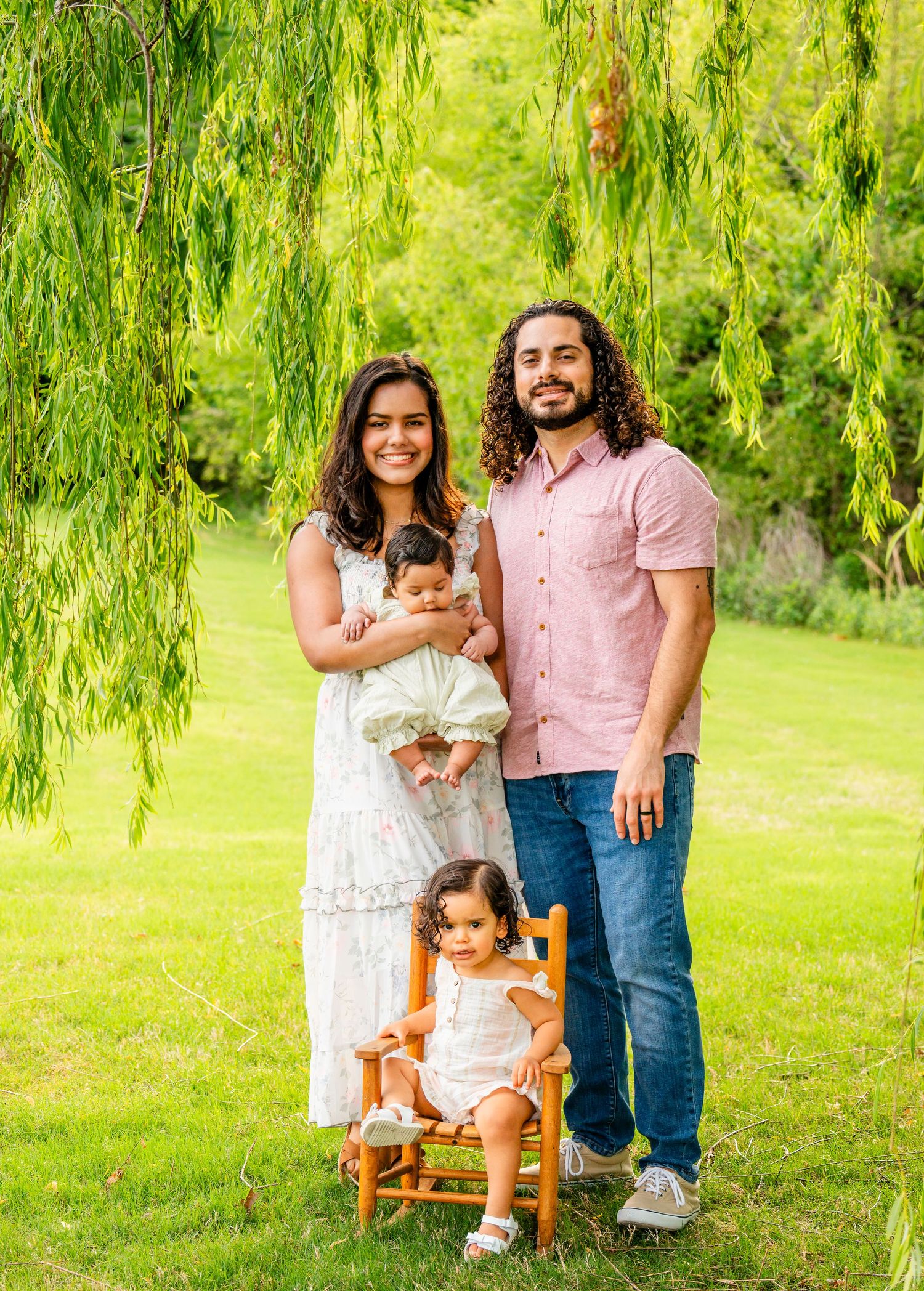 Family of four posing outdoors under a weeping willow tree, parents holding young children in a lush green park setting.
