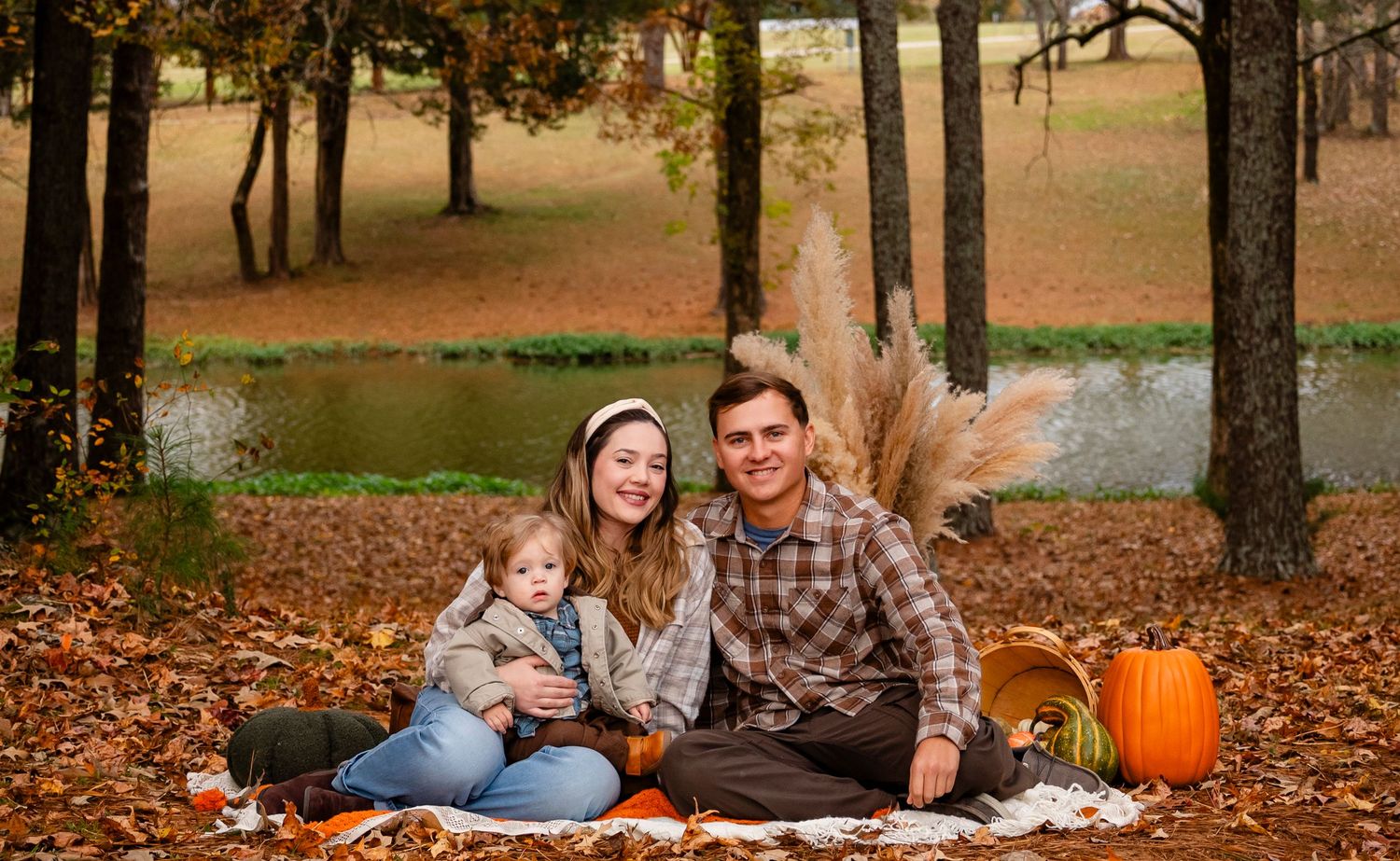 Happy family of three posing with pumpkins and fall decor on blanket in autumn leaves by pond.
