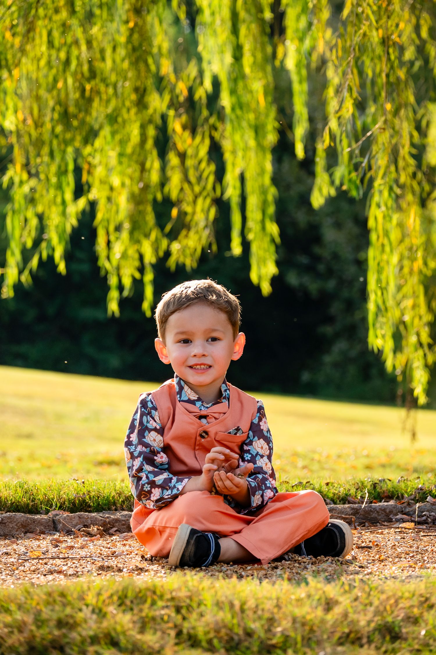 Young boy in orange pants and floral vest sitting cross-legged under a weeping willow tree outdoors.