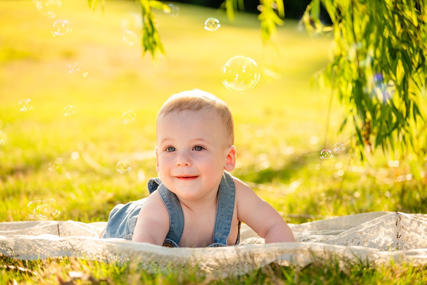 A smiling baby boy in denim overalls lying on a blanket outdoors surrounded by soap bubbles in sunny green grass.