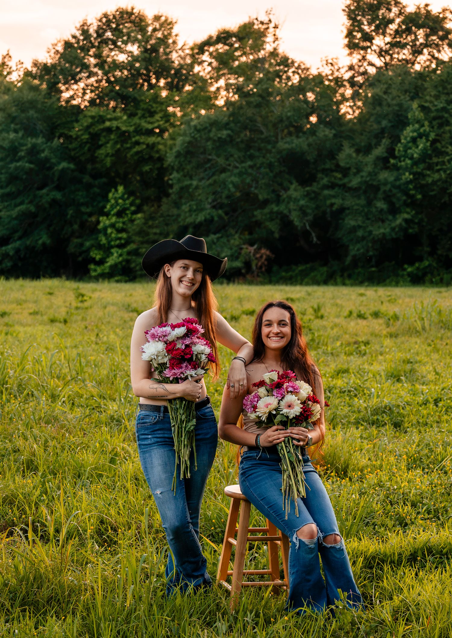 Two women holding flower bouquets posing in green field at sunset, one wearing cowboy hat, other seated on stool.