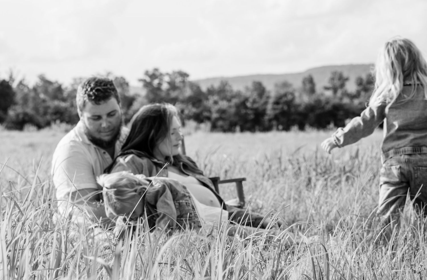 Black and white photo of a couple sitting in a field with a child playing nearby.