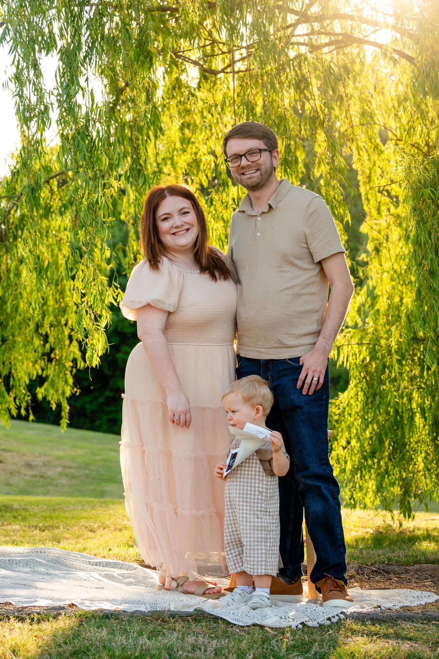 Family of three posing under weeping willow tree in warm golden hour sunlight outdoors.