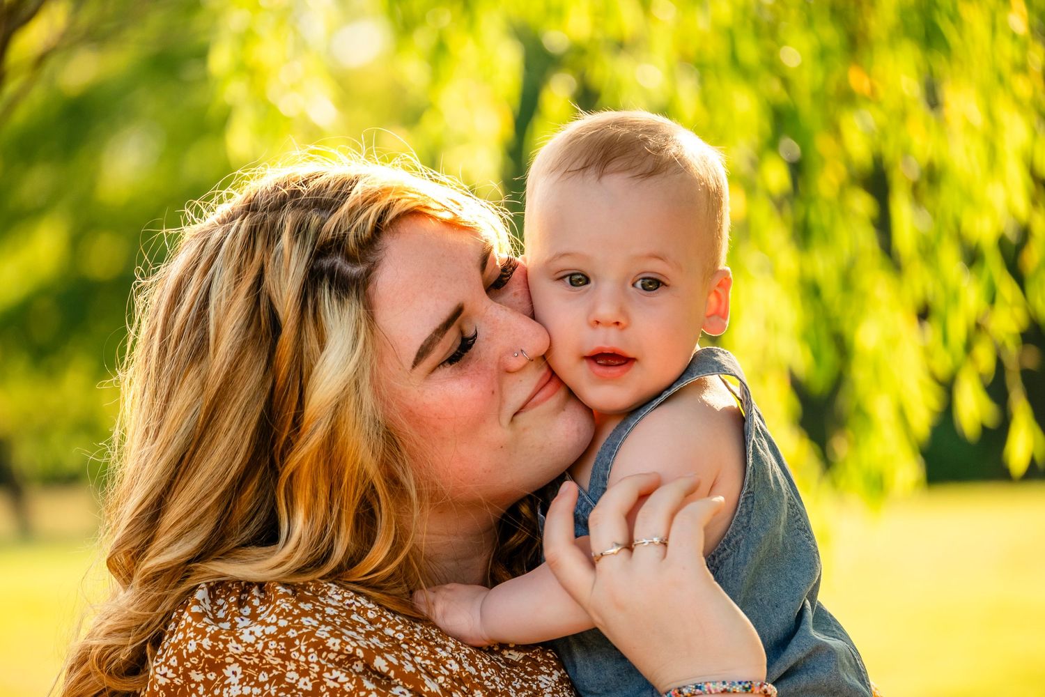 A mother tenderly kisses her baby boy on the cheek outdoors in a sunny green park setting.