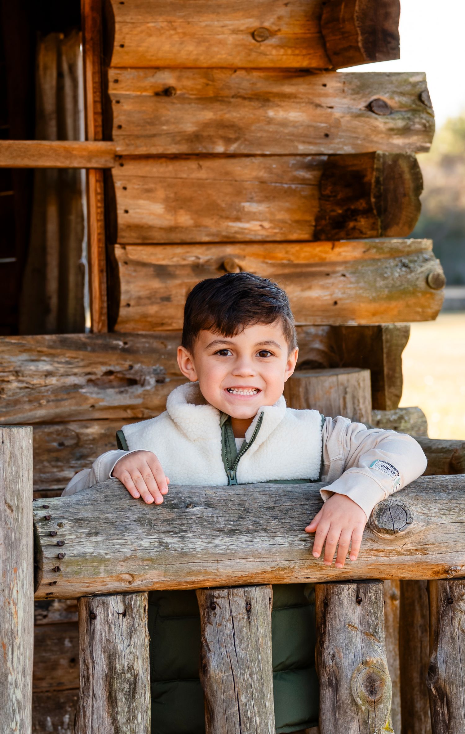 A smiling young Hispanic boy leans on a rustic log cabin fence railing in warm afternoon light.