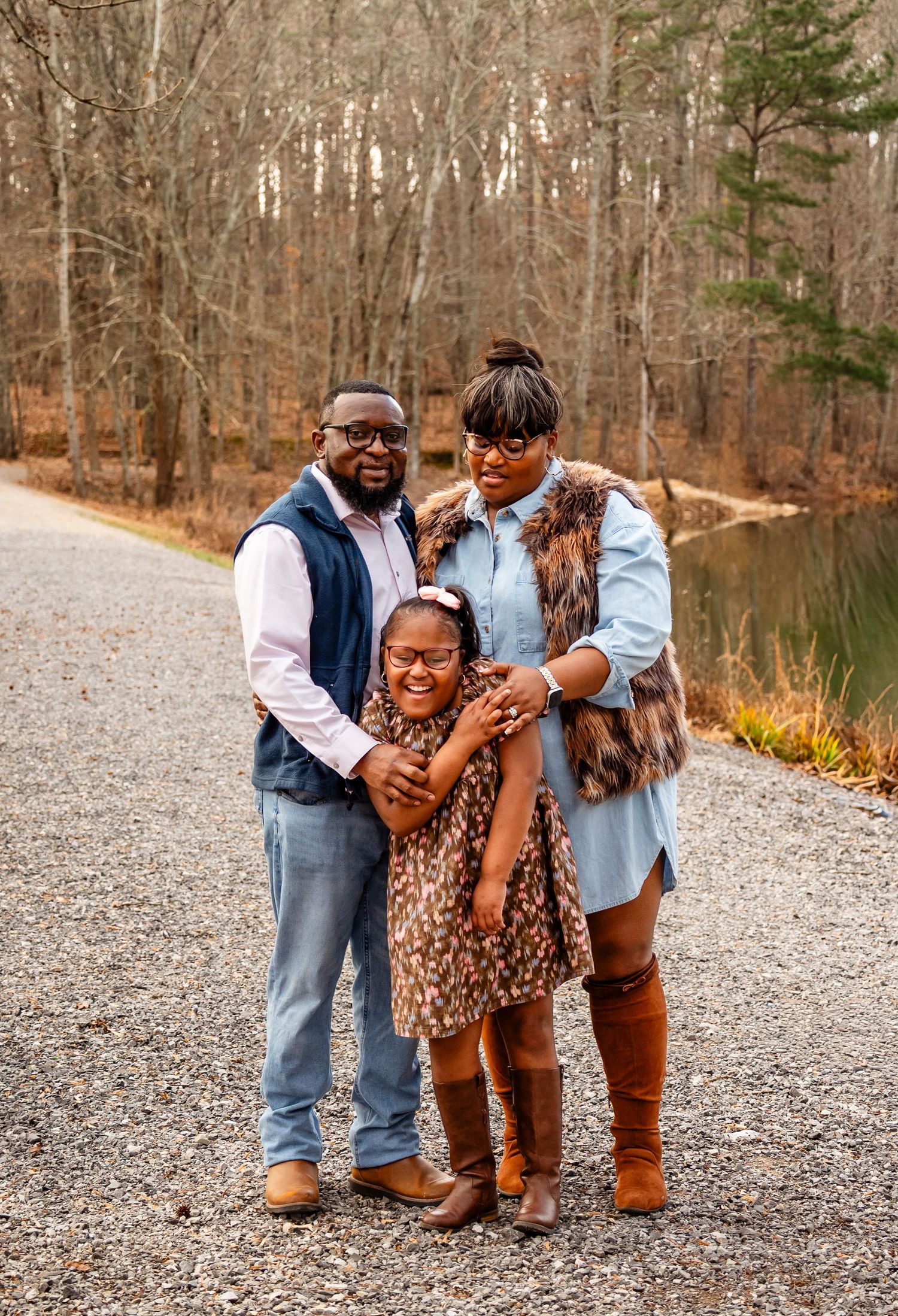 A Black family of three poses on a gravel path surrounded by bare autumn trees near a pond.