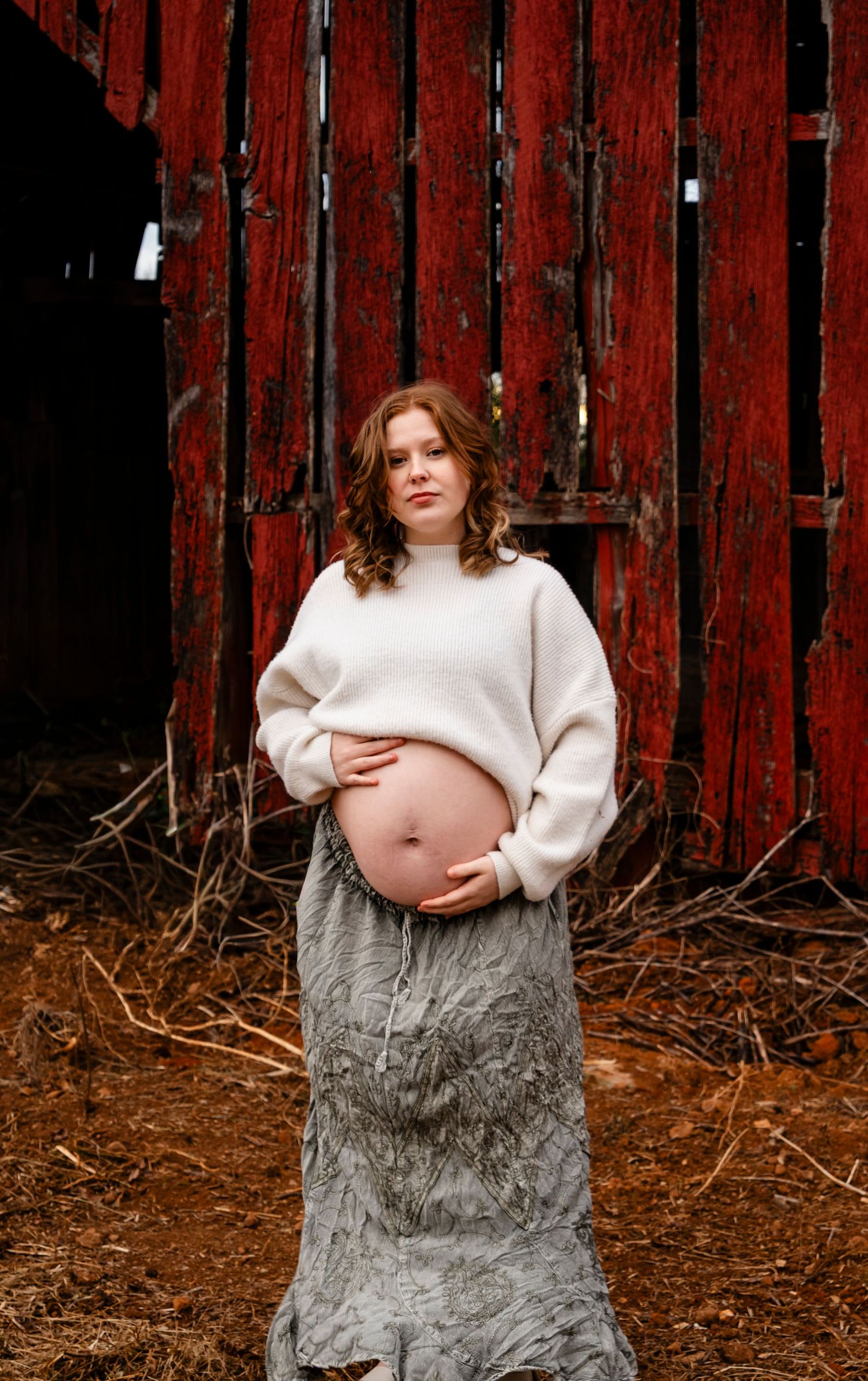 Expectant mother in cream sweater holding pregnant belly while standing before a weathered red barn door.