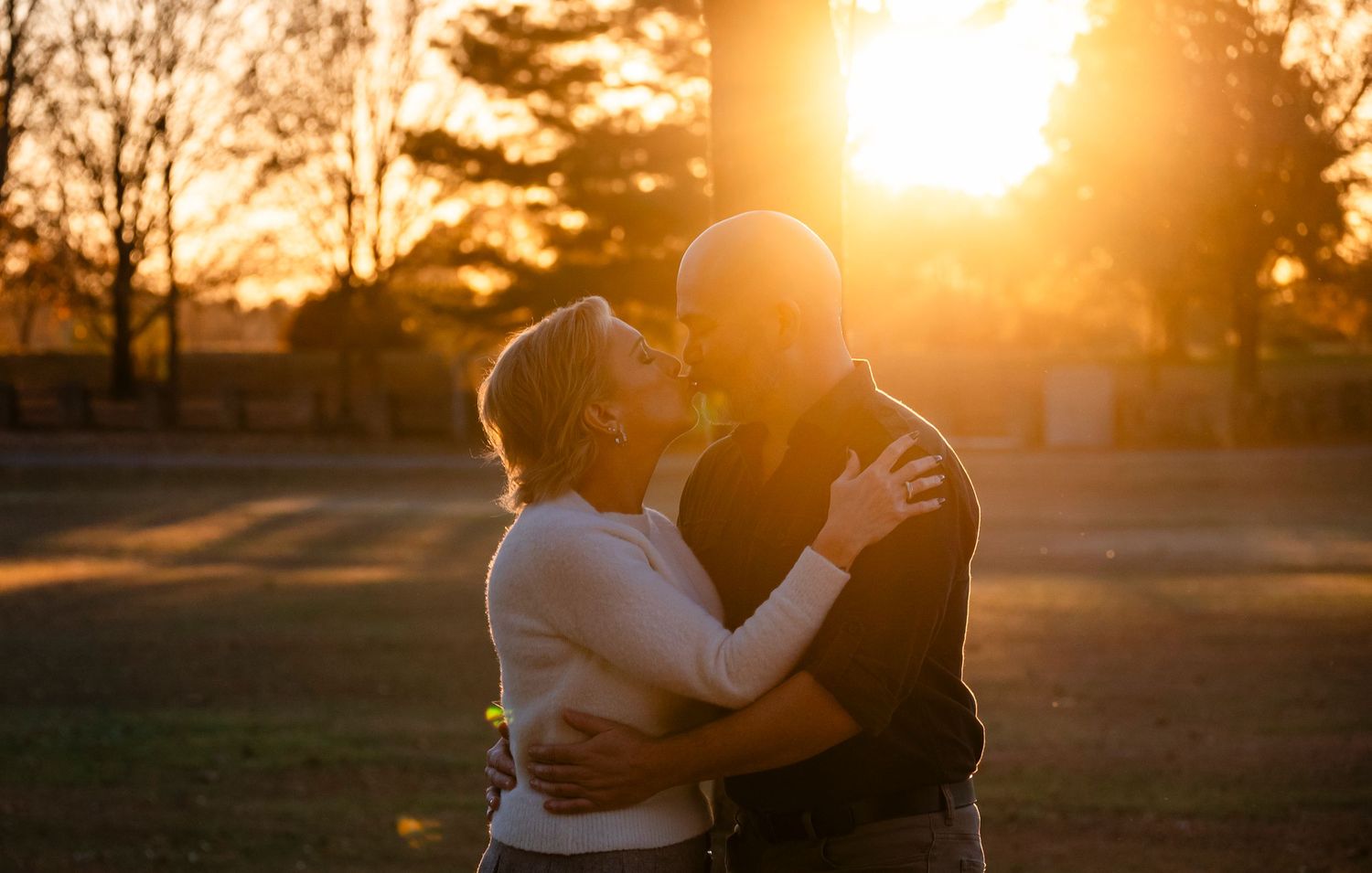 Couple sharing a kiss at golden sunset outdoors, silhouetted against warm glowing light in an open field.