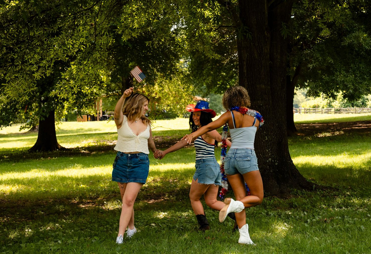 Three young women dancing and playing in a lush green park under large shade trees on a sunny day.