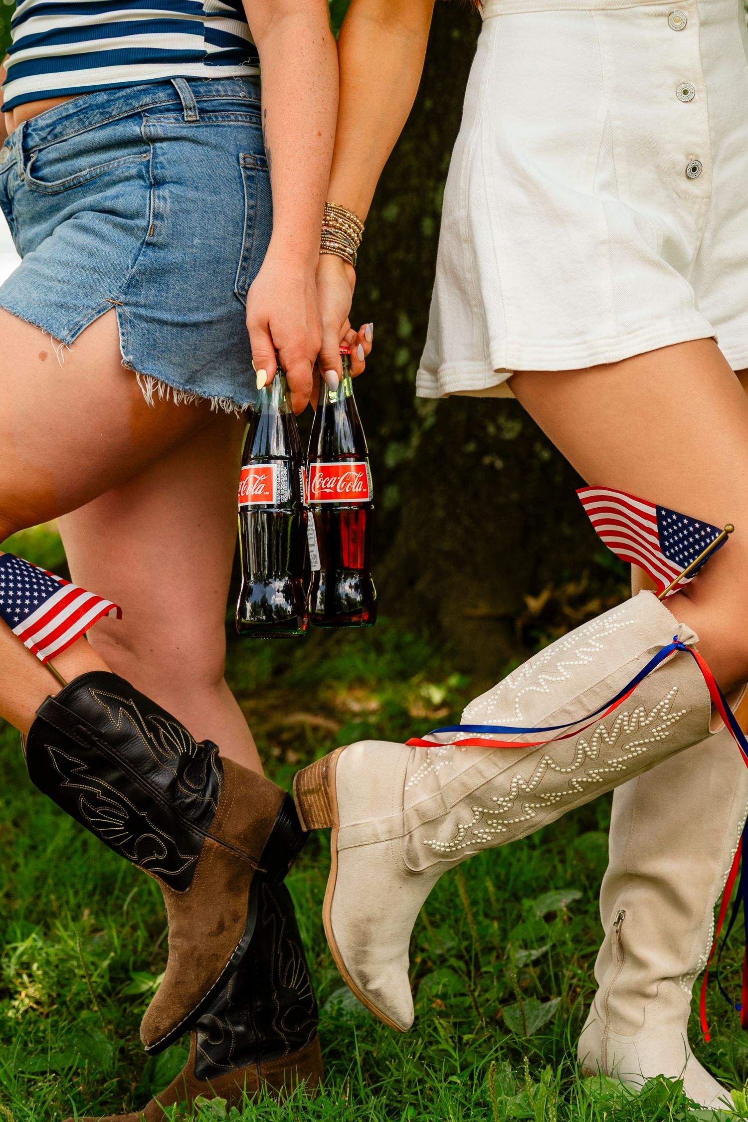 Two women in denim shorts and cowboy boots hold Coca-Cola bottles with American flags tucked into their boots.