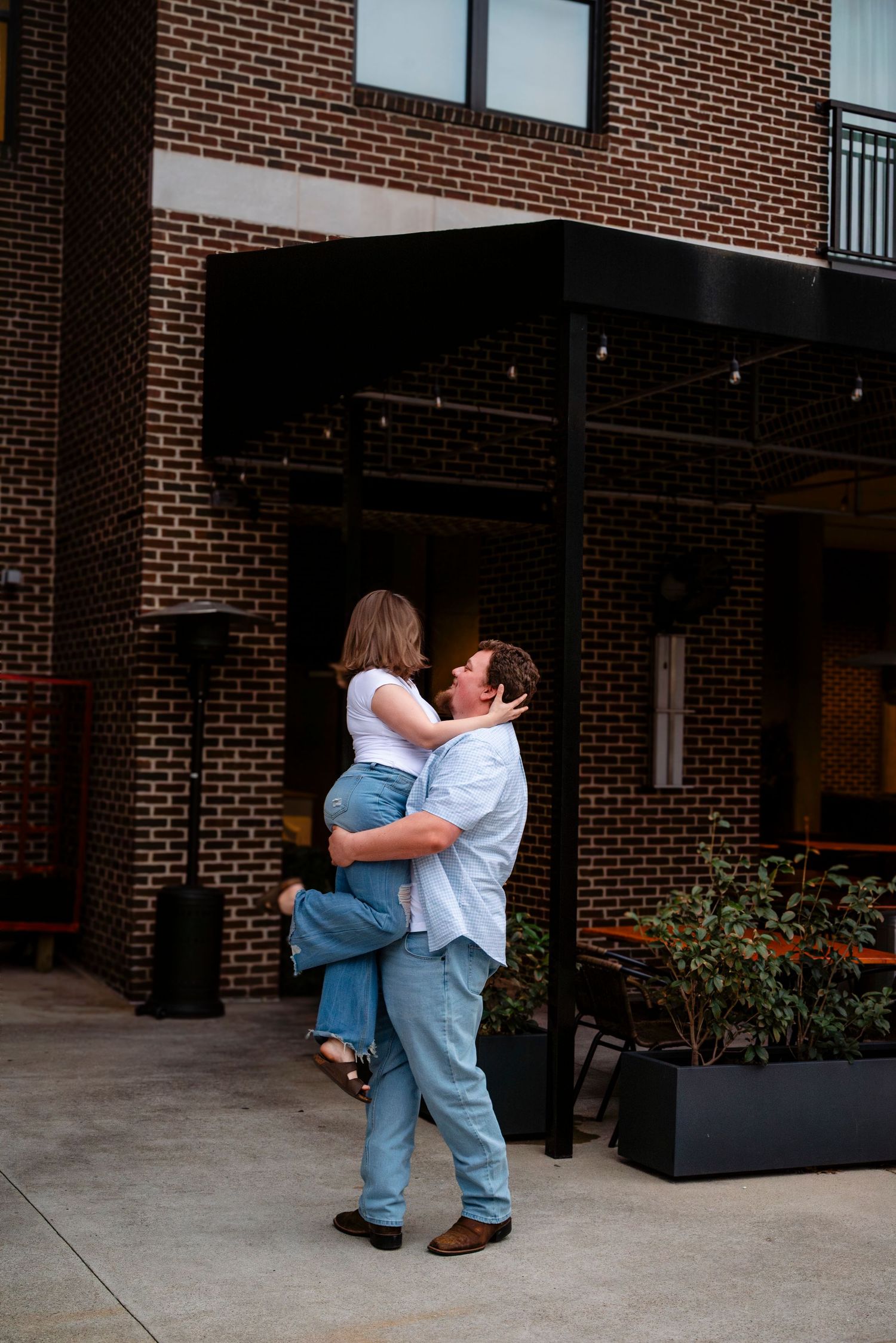 A man lifts a woman in his arms outside a brick building patio, sharing a romantic moment.