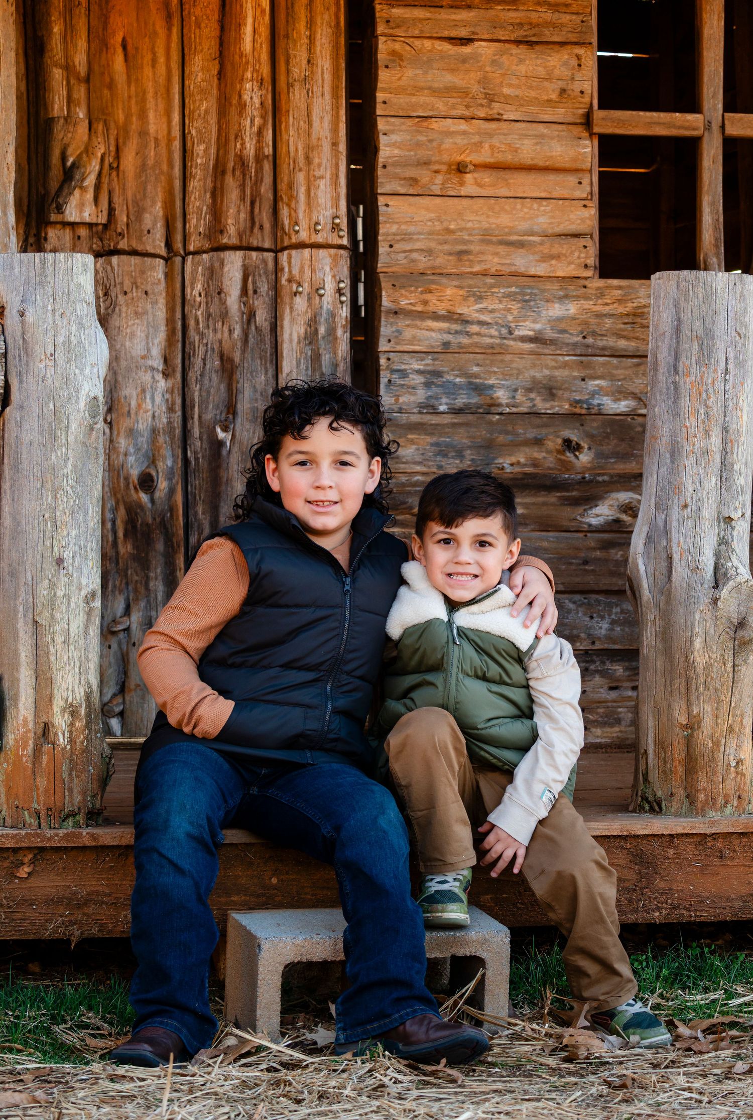Two young Hispanic brothers sit together smiling in front of a rustic wooden barn wall.