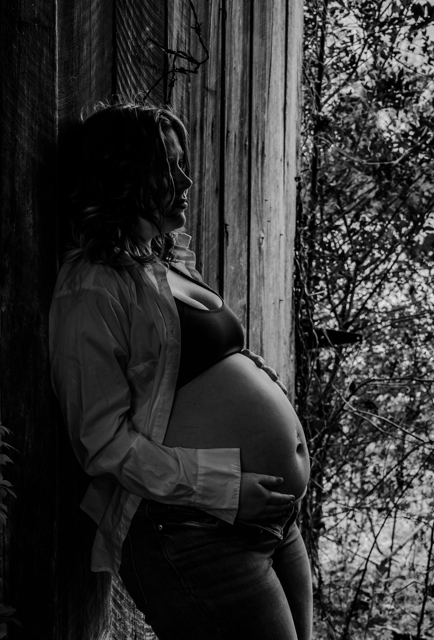 Side profile of pregnant woman leaning against weathered wood wall, cradling baby bump, black and white maternity portrait.