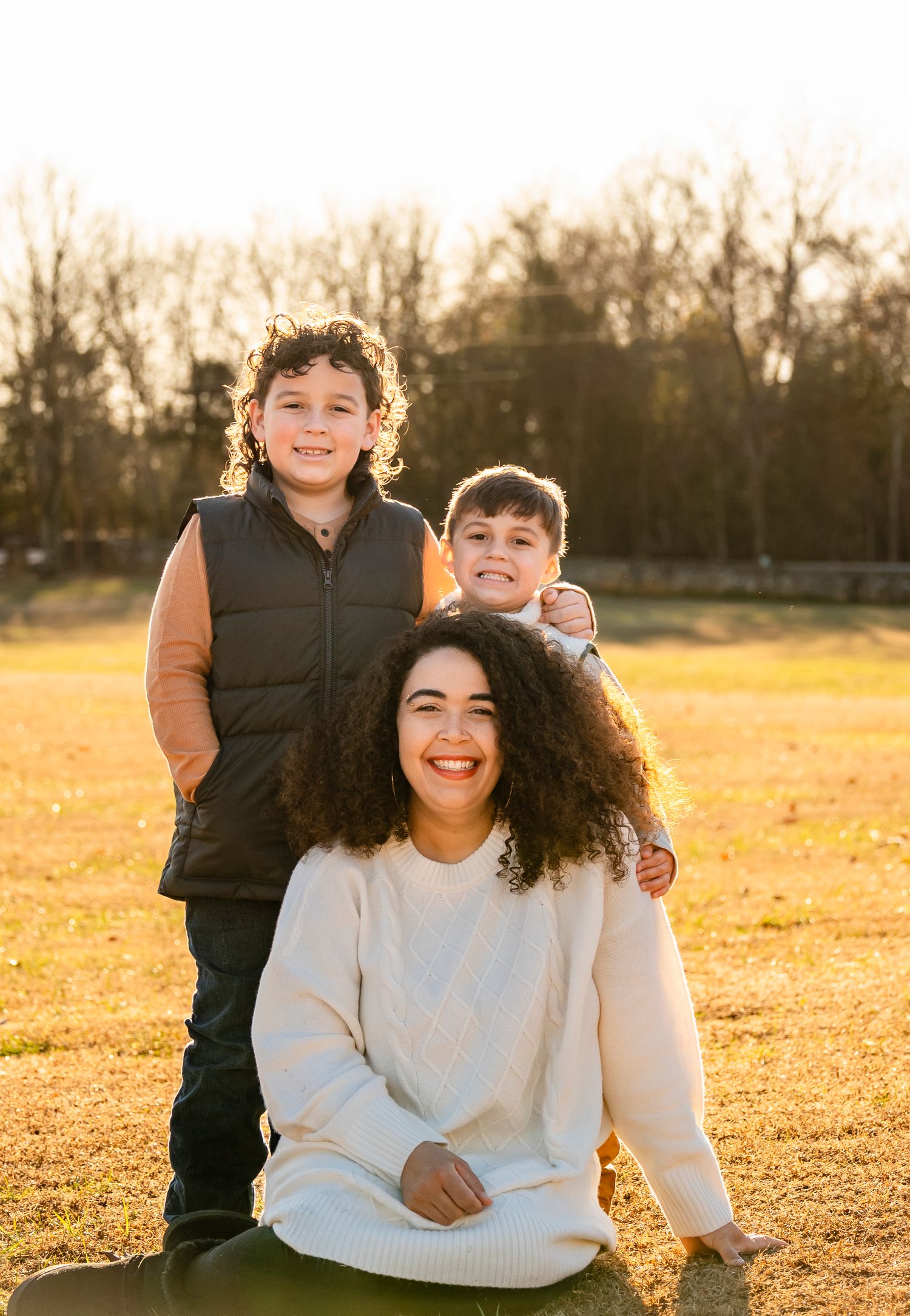 A laughing curly-haired mother poses outdoors with her two boys standing behind her at sunset.