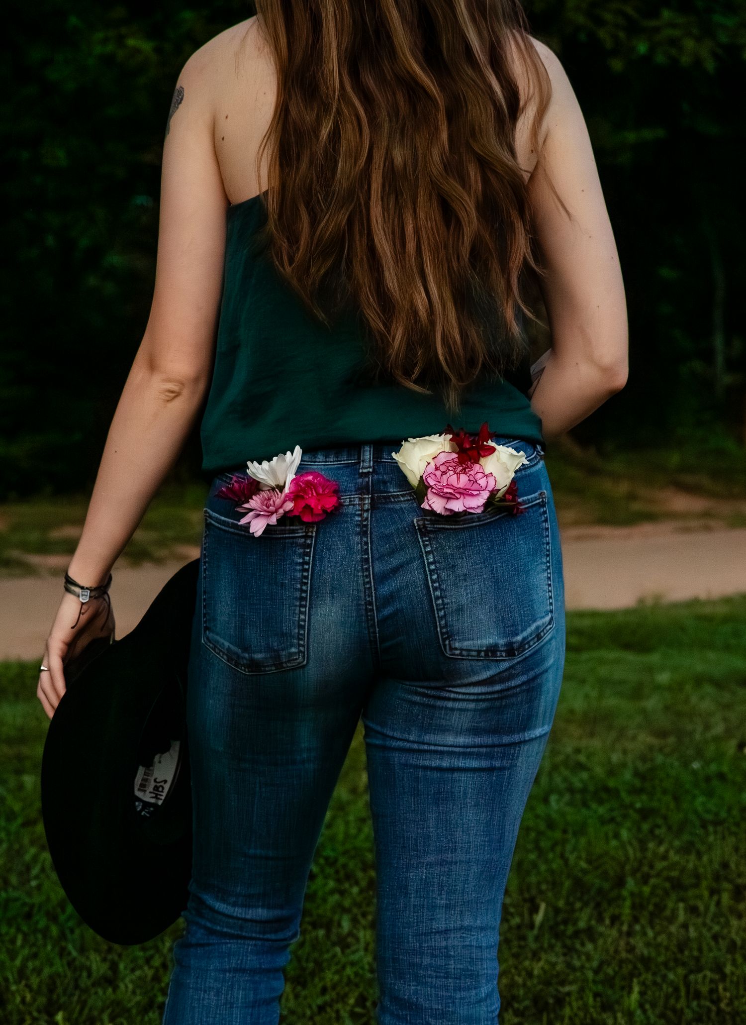 Back view of a woman in green top and jeans with colorful flowers tucked in back pockets, holding a black hat.
