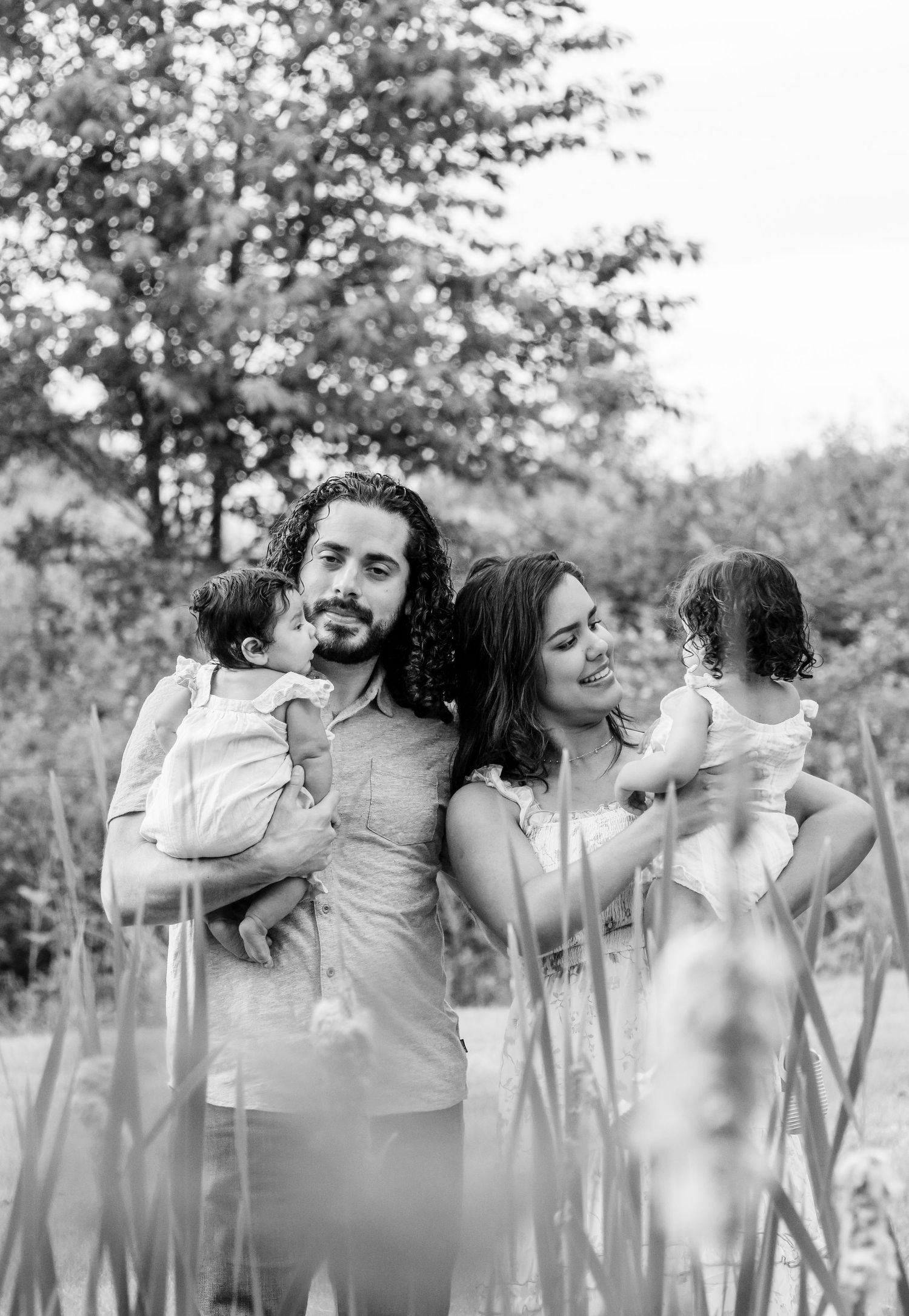 Black and white family portrait of parents holding two young children outdoors among tall reeds with trees in background.