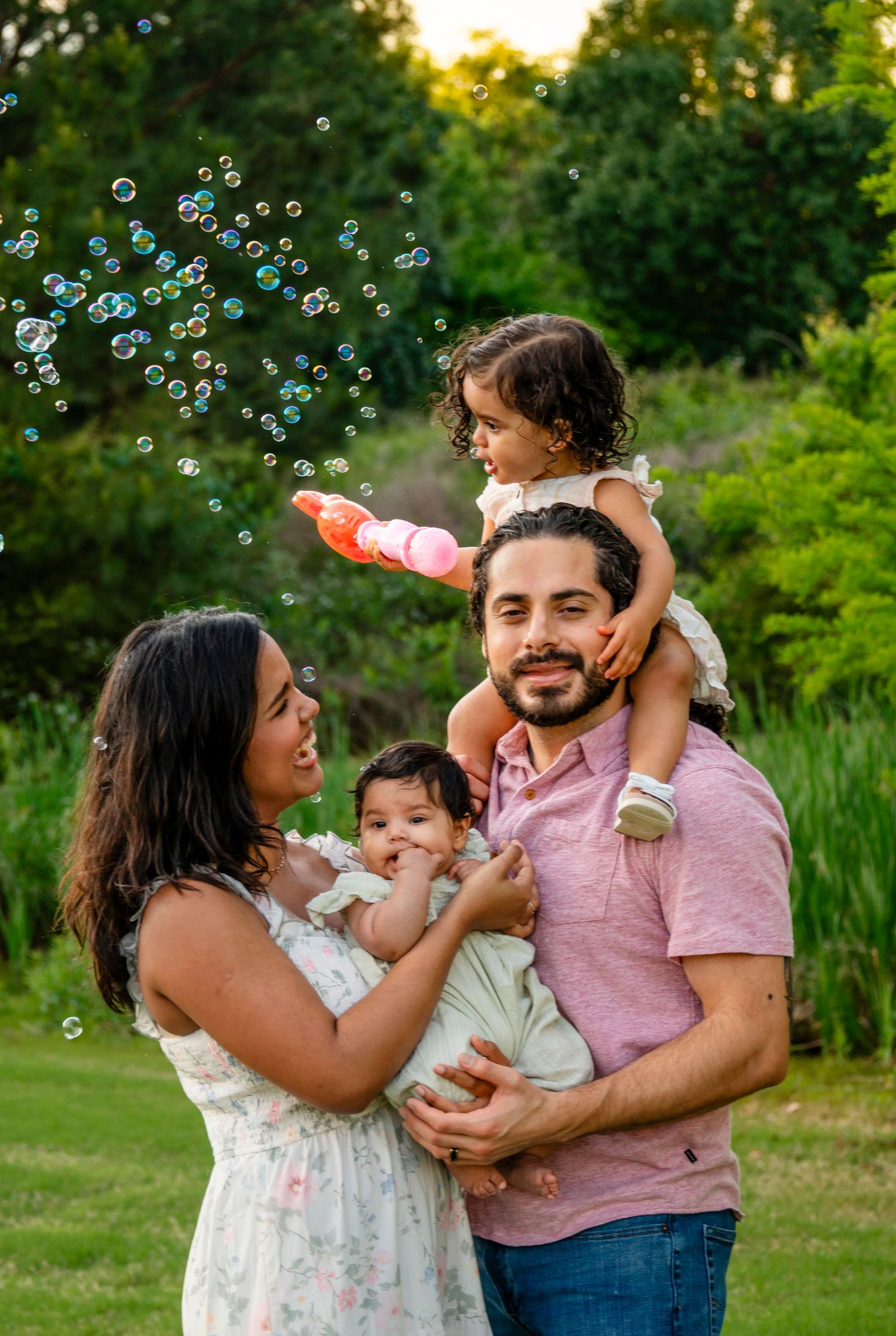 A happy family with two young children enjoy bubbles together in a lush green park at sunset.