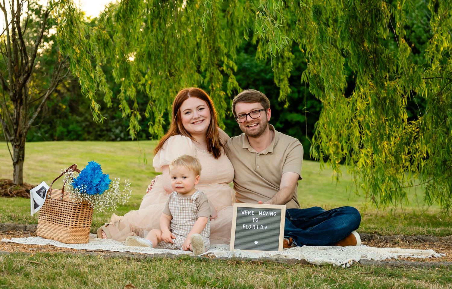 Expecting family sits on blanket outdoors with toddler and letter board pregnancy announcement sign.