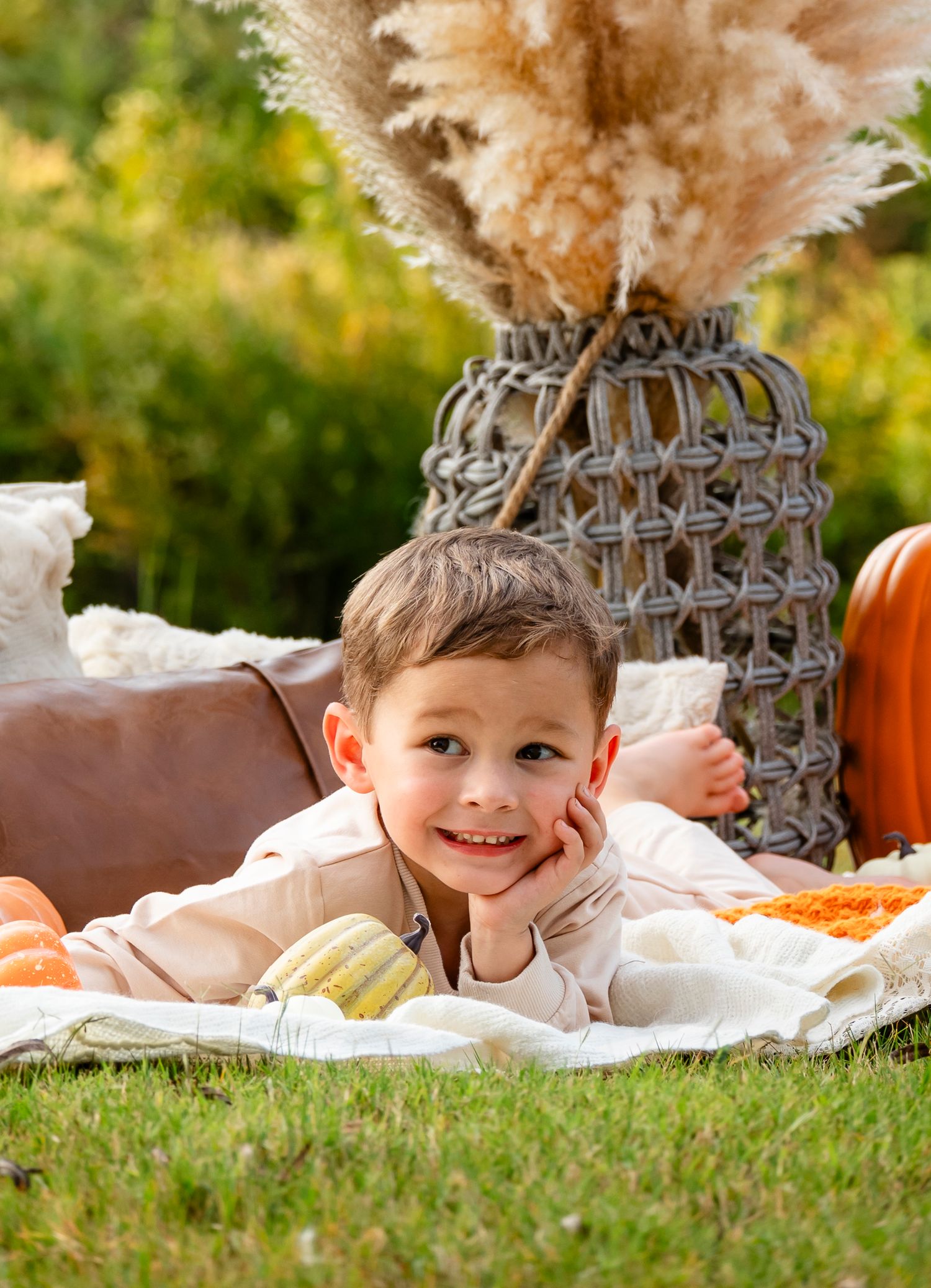 Smiling young boy lying on blanket with pumpkins and pampas grass in autumn outdoor setting.
