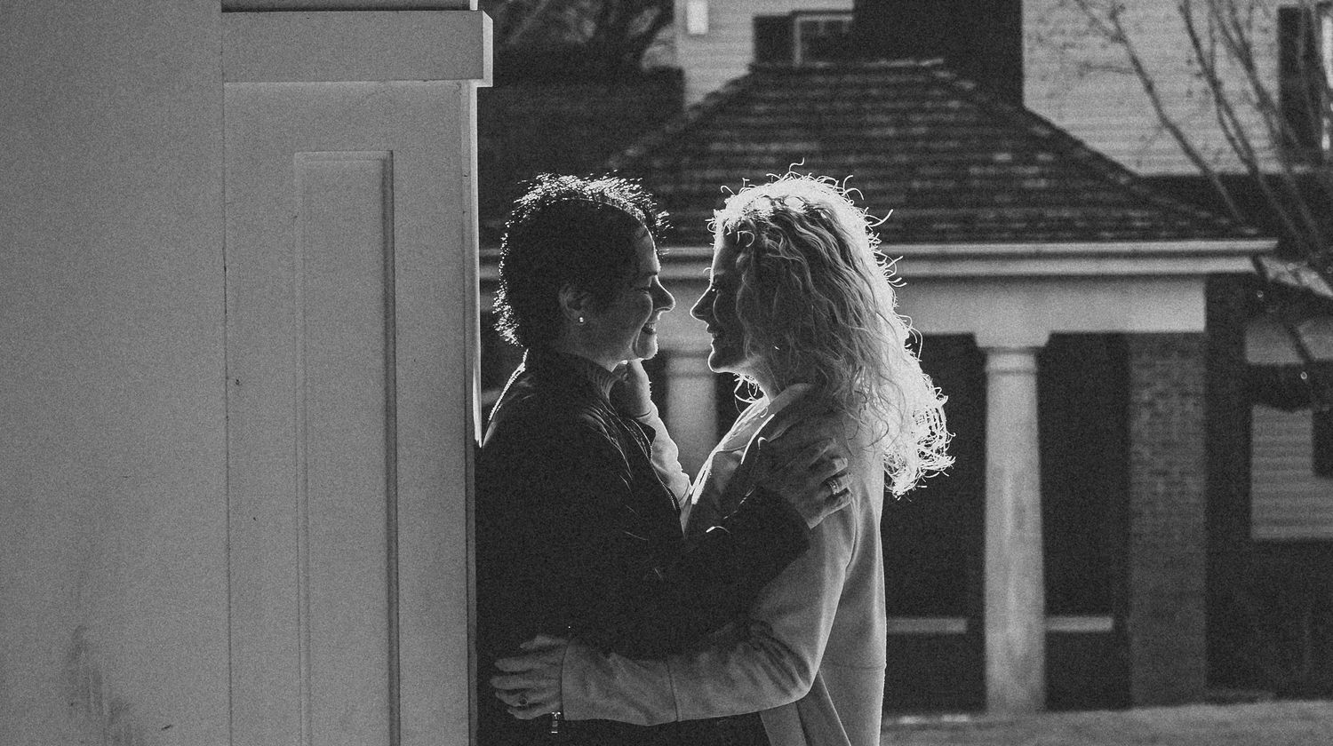 Couple sharing an intimate moment under a white column with a house visible in the background.