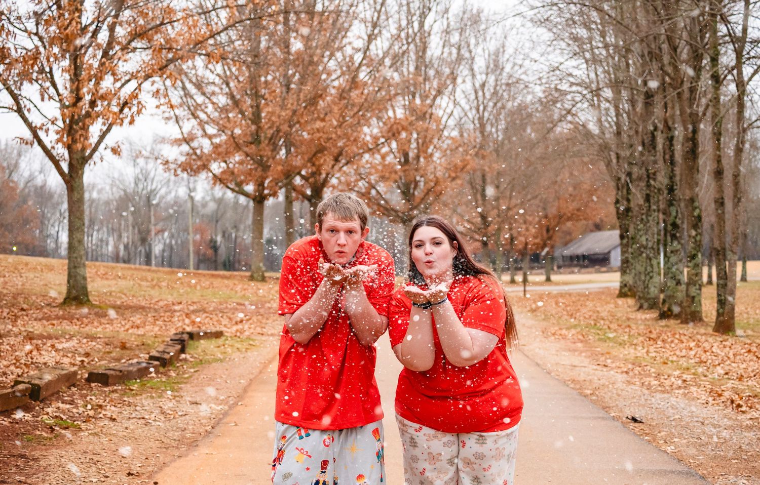 Couple in matching red Christmas pajamas blowing fake snow outdoors among fall foliage trees.