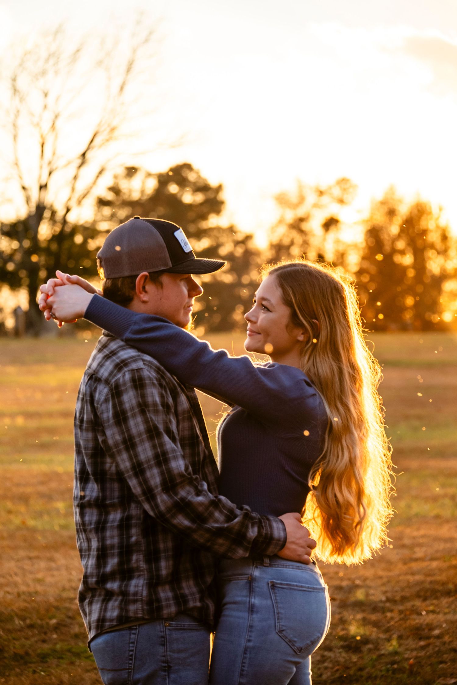 Couple embracing at golden hour sunset in a field, woman smiling with long hair glowing in warm light.