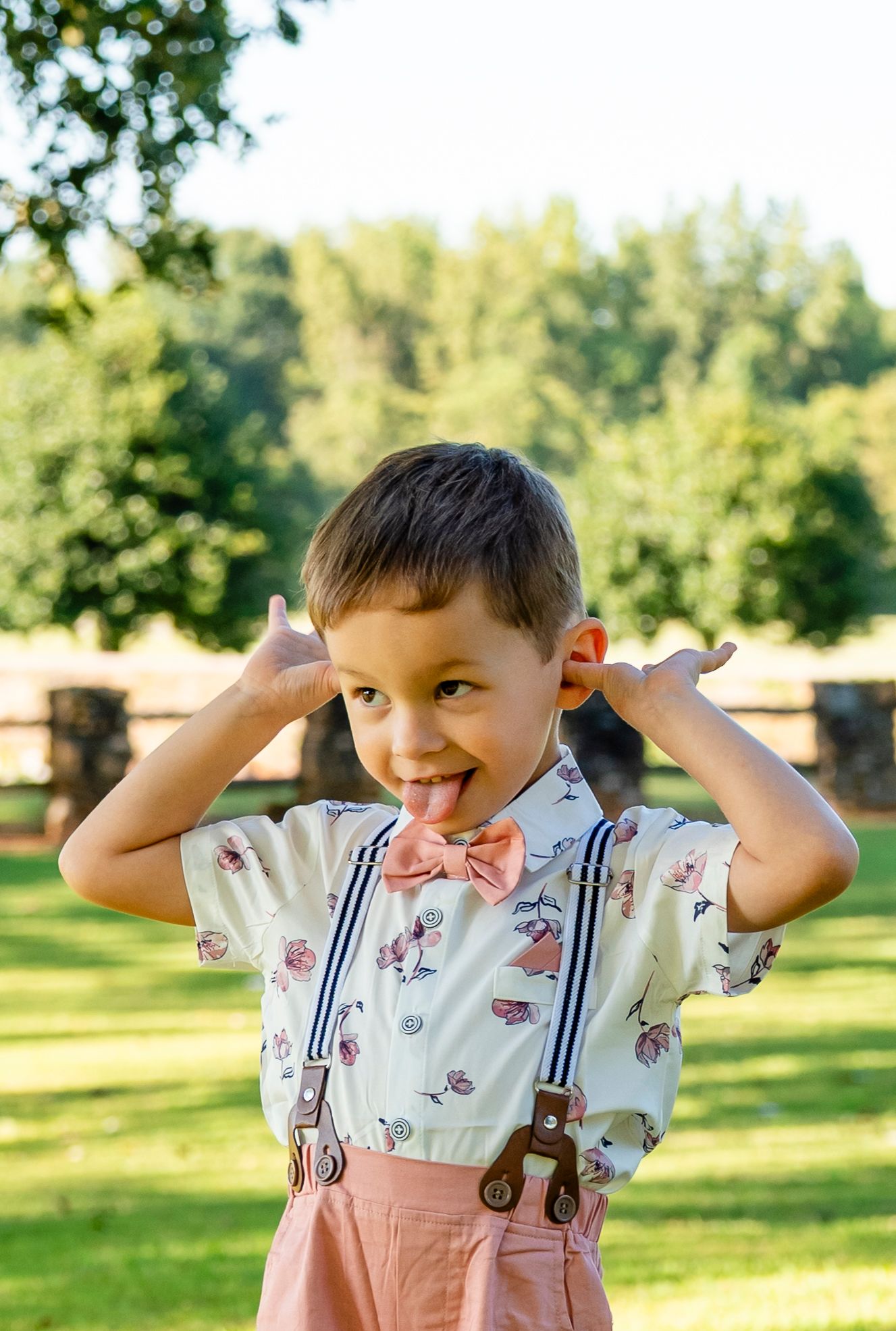 Funny young boy in bow tie and suspenders making silly face with fingers on ears and tongue out in park.