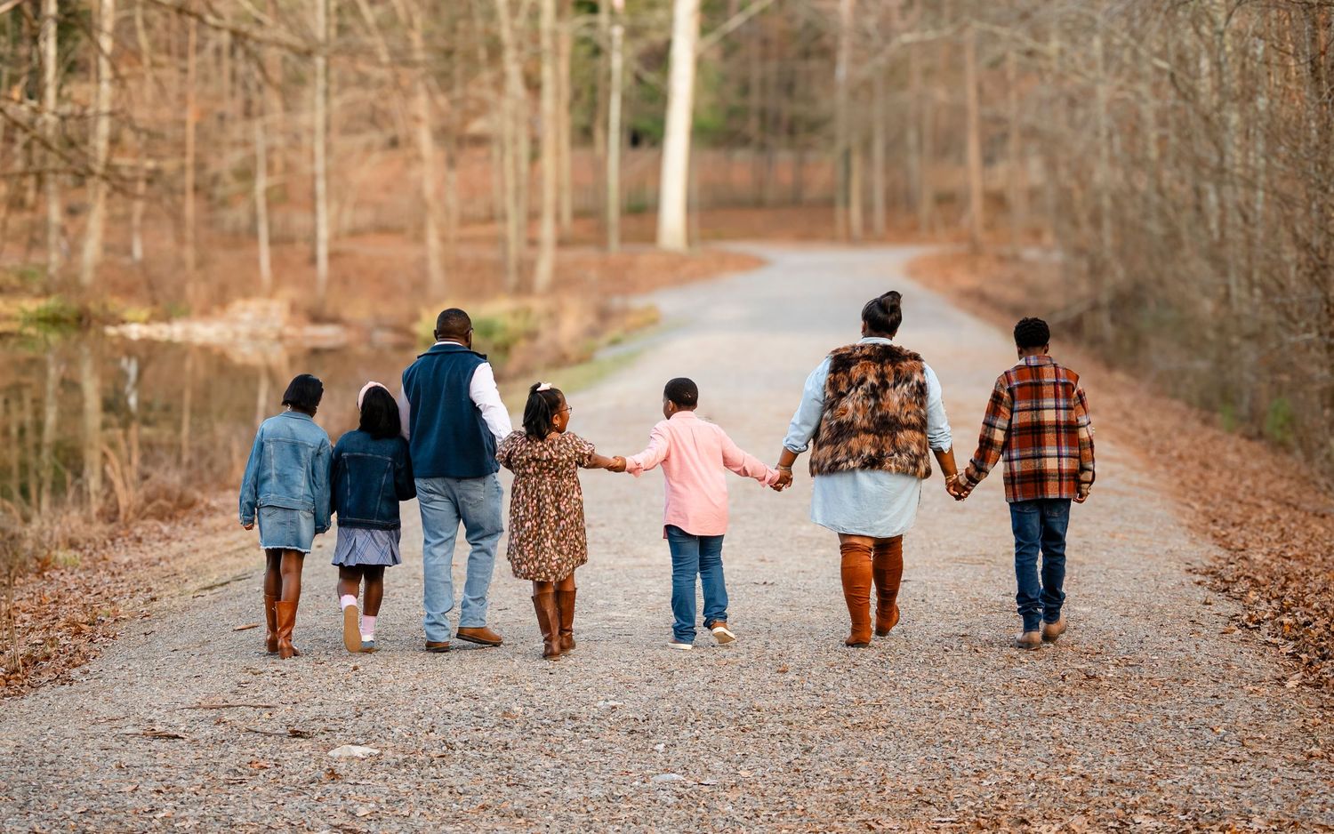 A family of seven walks hand-in-hand down a scenic autumn tree-lined gravel path from behind.