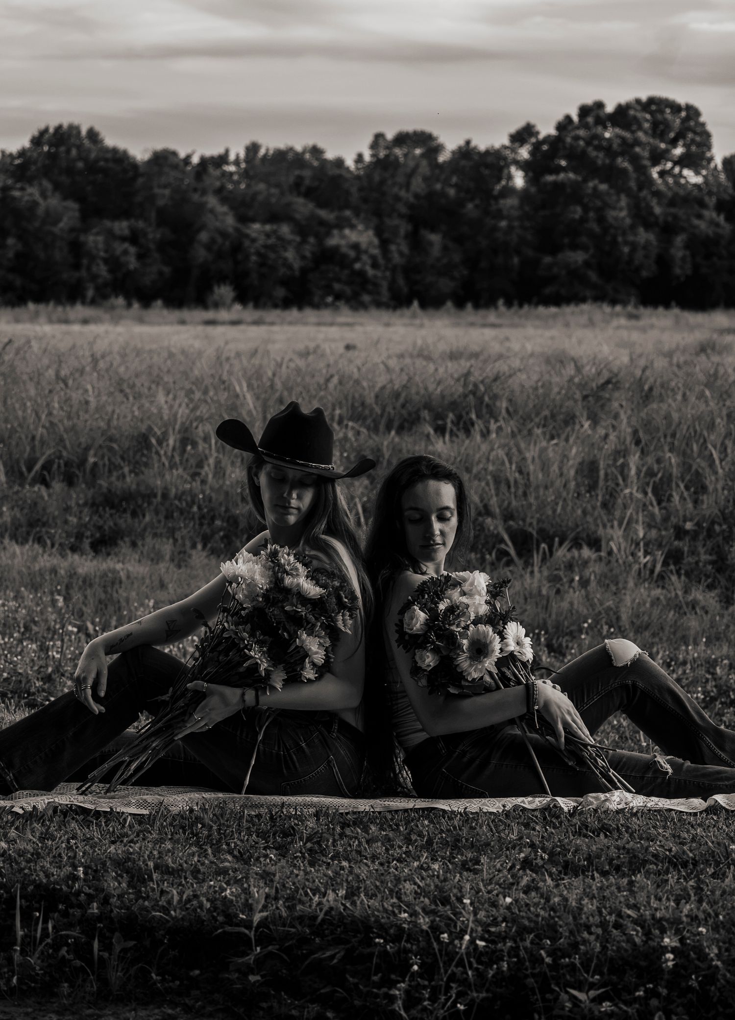 Two women sit back-to-back in a field holding flower bouquets, one wearing a cowboy hat, in black and white.