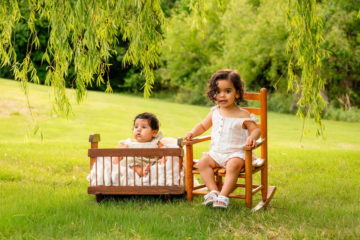 Two young children pose outdoors, one in a wooden cradle and one in a rocking chair, on a lush green lawn.