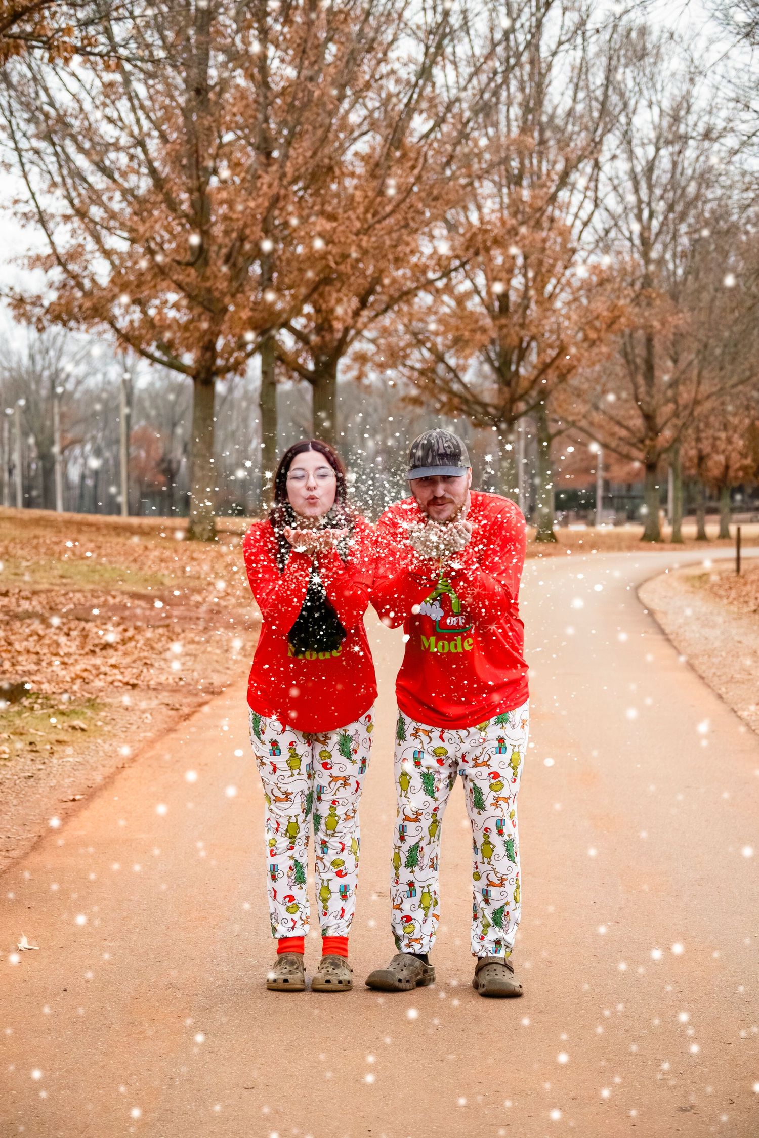 Couple in matching red Grinch pajamas blow glitter on a tree-lined path in a snowy setting.